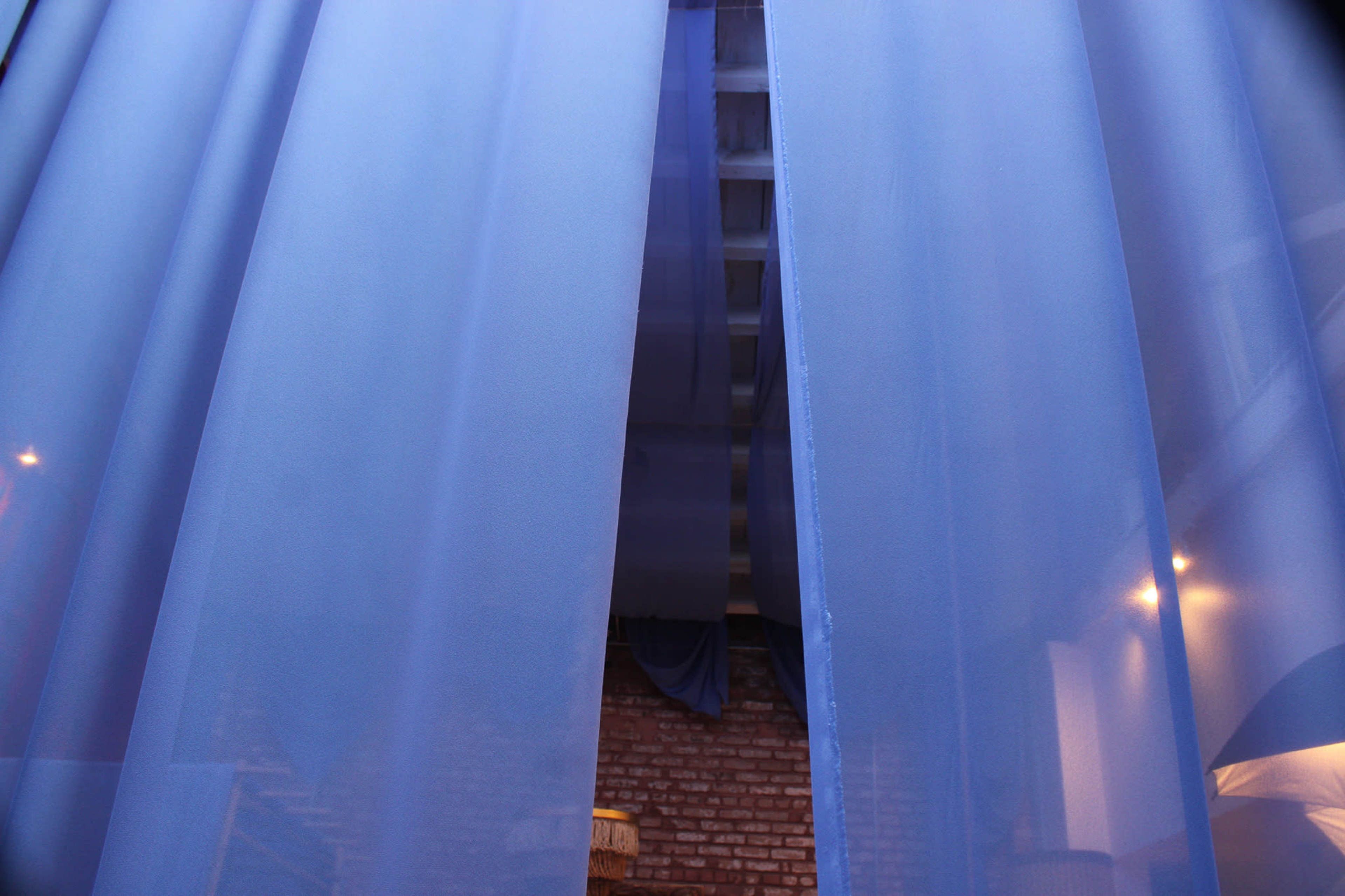 A view through sheer blue curtains into a softly lit interior space with exposed brick walls and a staircase.