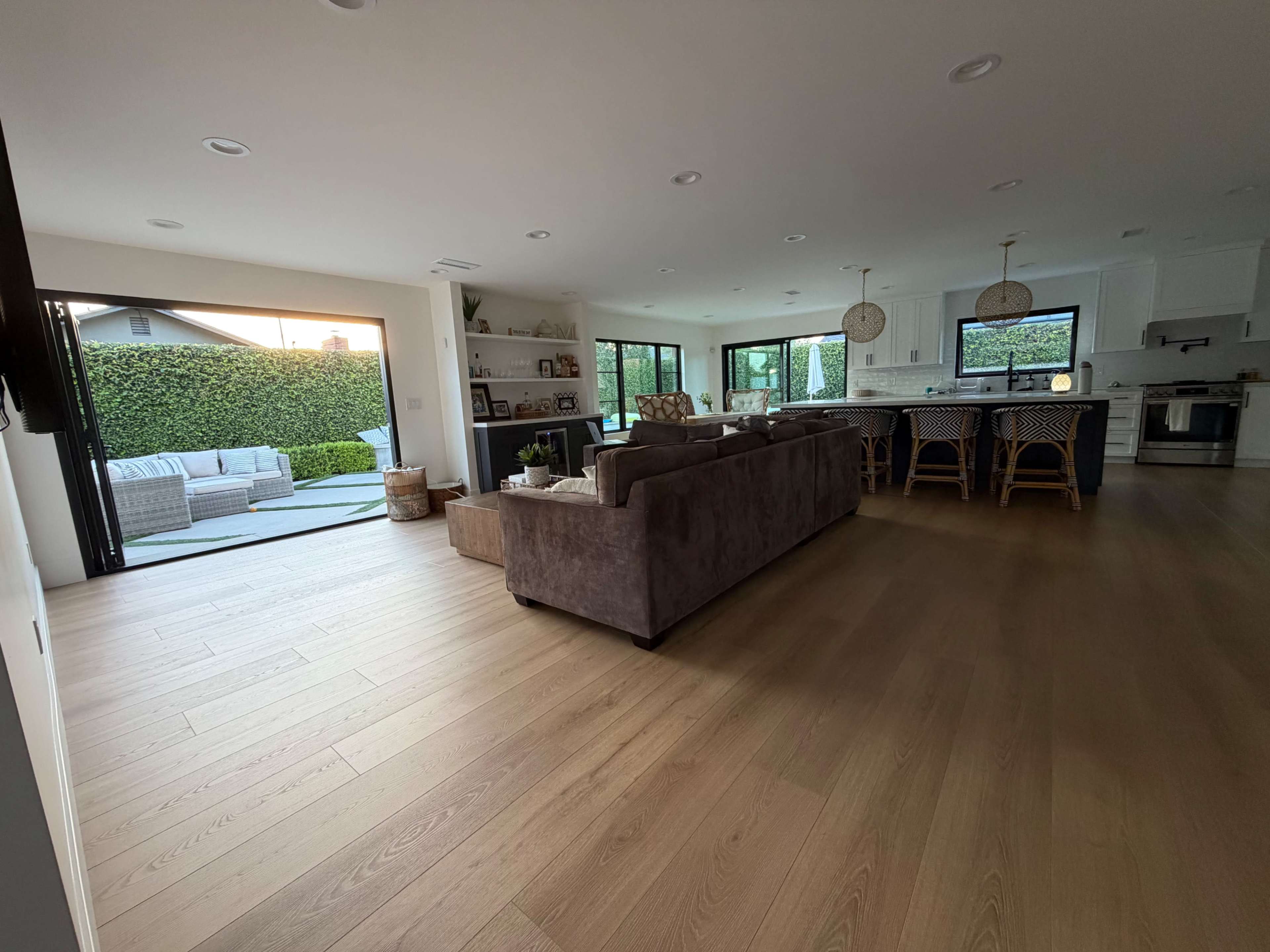 The image shows a modern open-concept living area with a view of a patio through large glass doors, featuring a comfortable seating arrangement and a kitchen in the background.