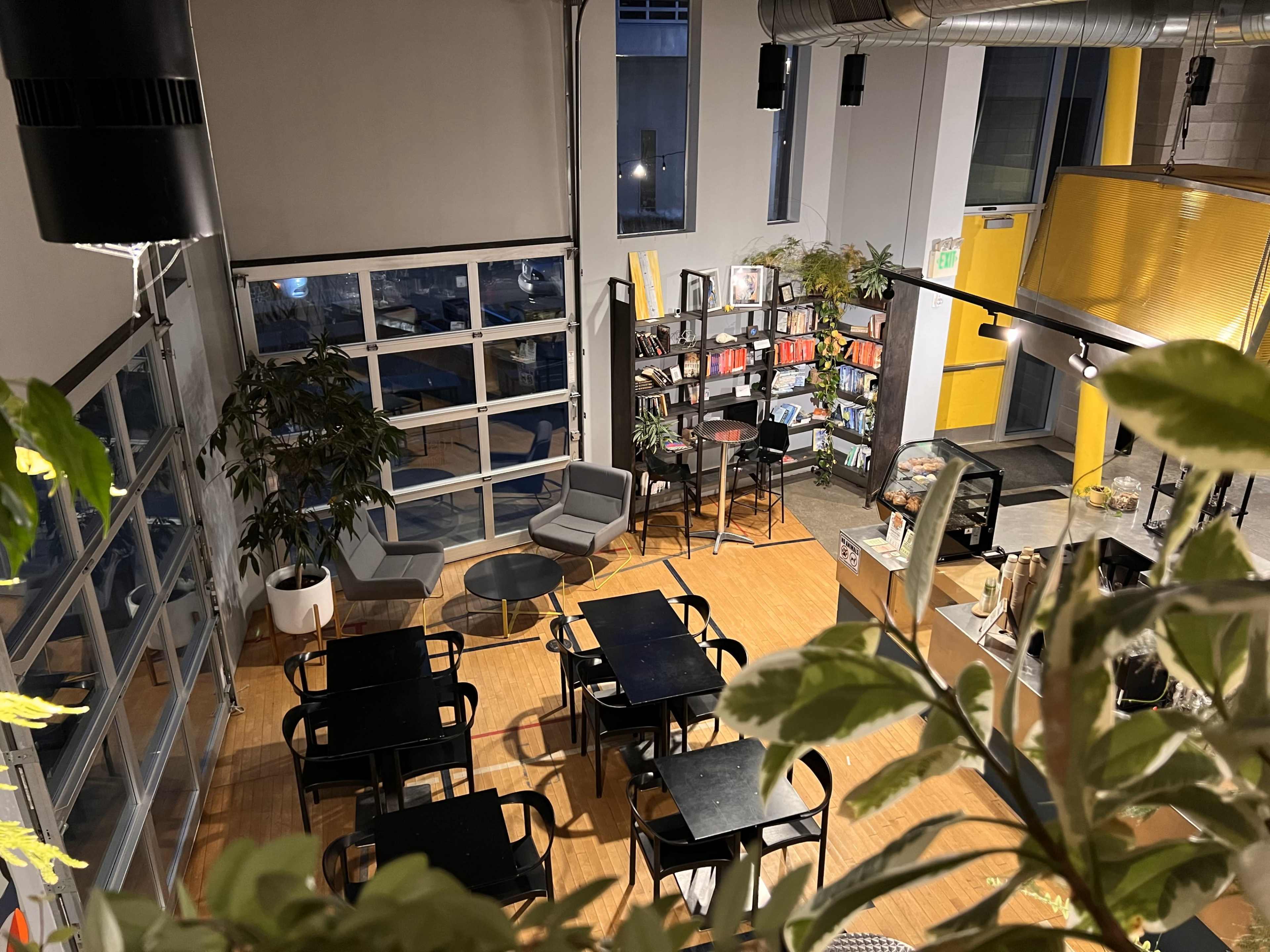 A modern café with several black tables and chairs, bookshelves, and large windows allowing natural light to enter.