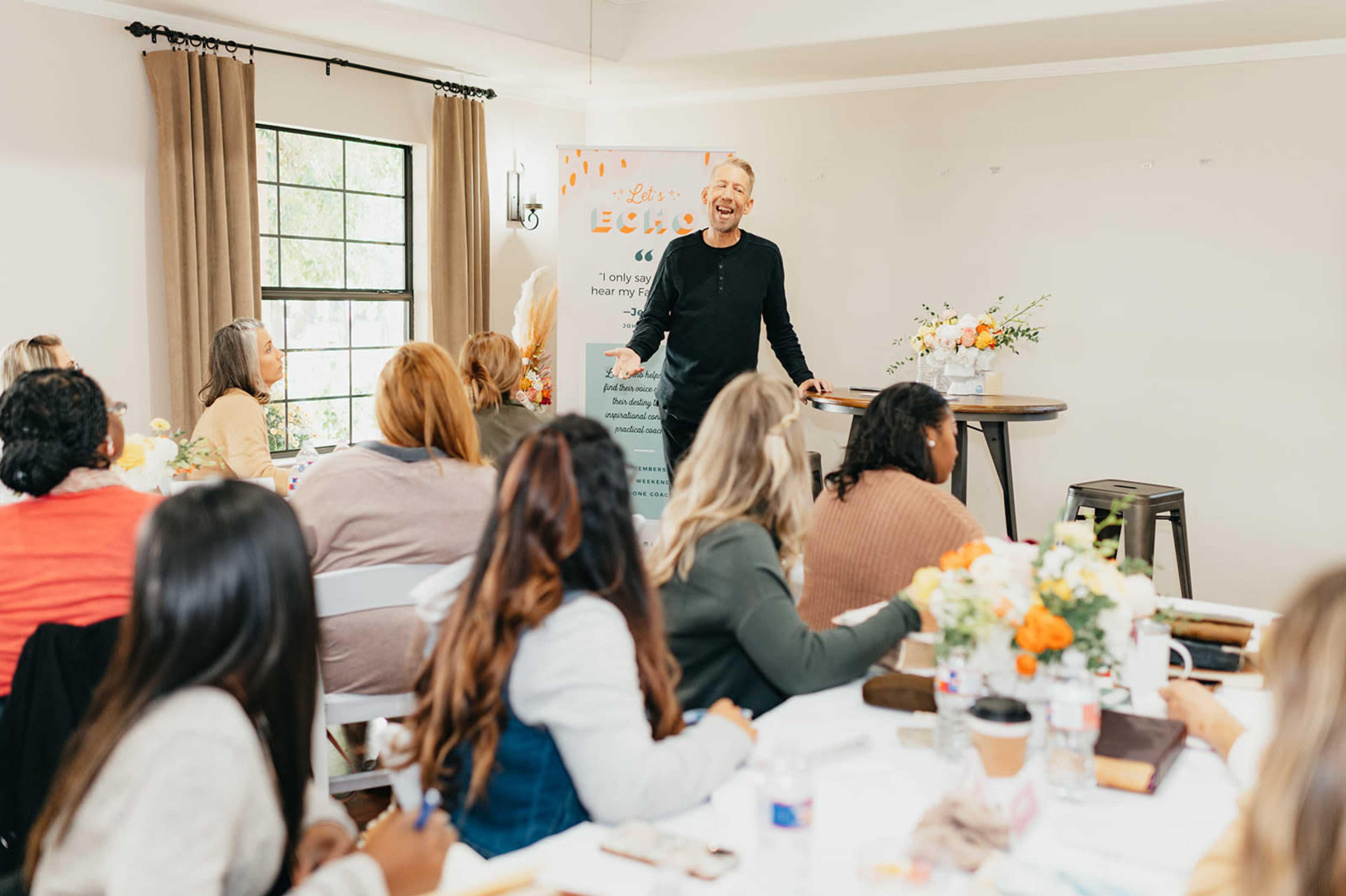 A speaker stands at the front of a well-lit room, engaging an audience seated at tables decorated with flowers and notebooks.