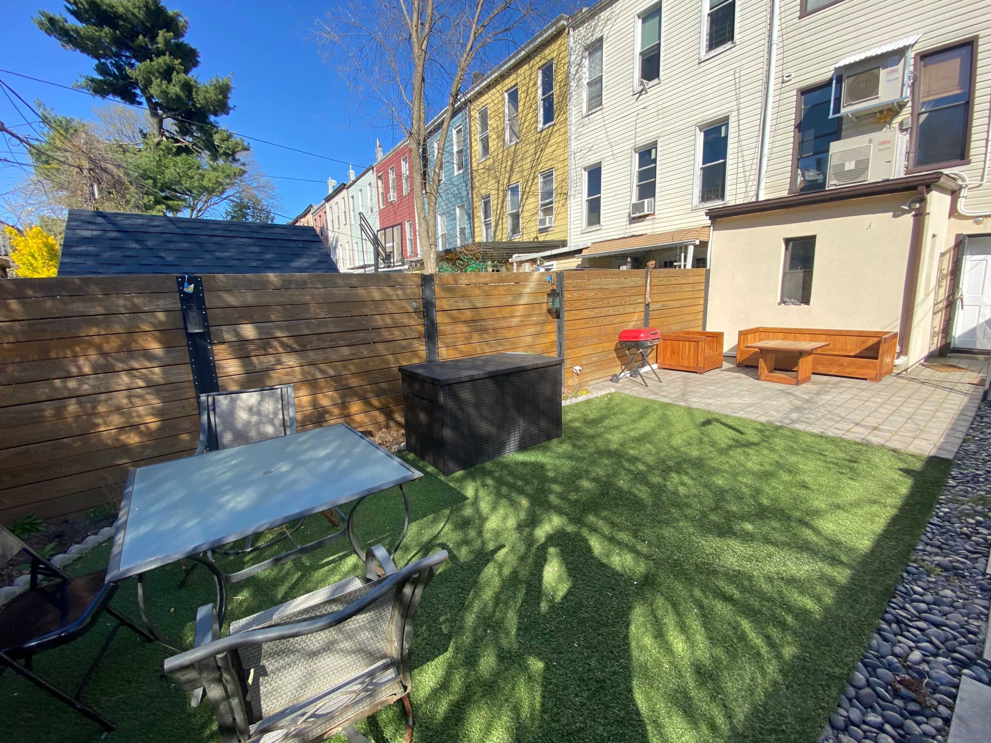 A backyard area with artificial grass, a small table and chairs, a storage box, a grill, and neighboring houses in the background.