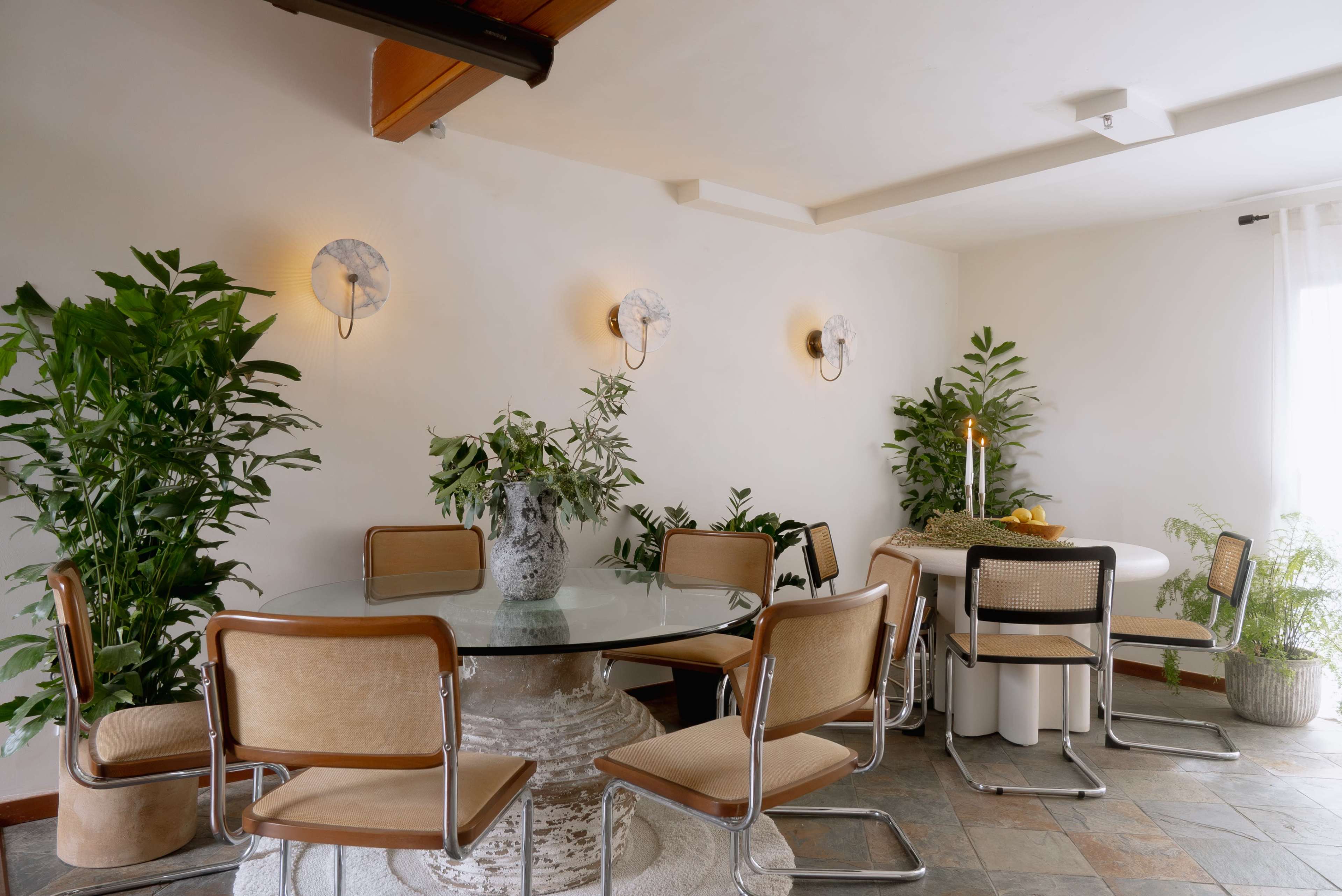 A modern dining area features a circular glass table surrounded by upholstered chairs, with greenery in decorative pots and wall-mounted lighting.