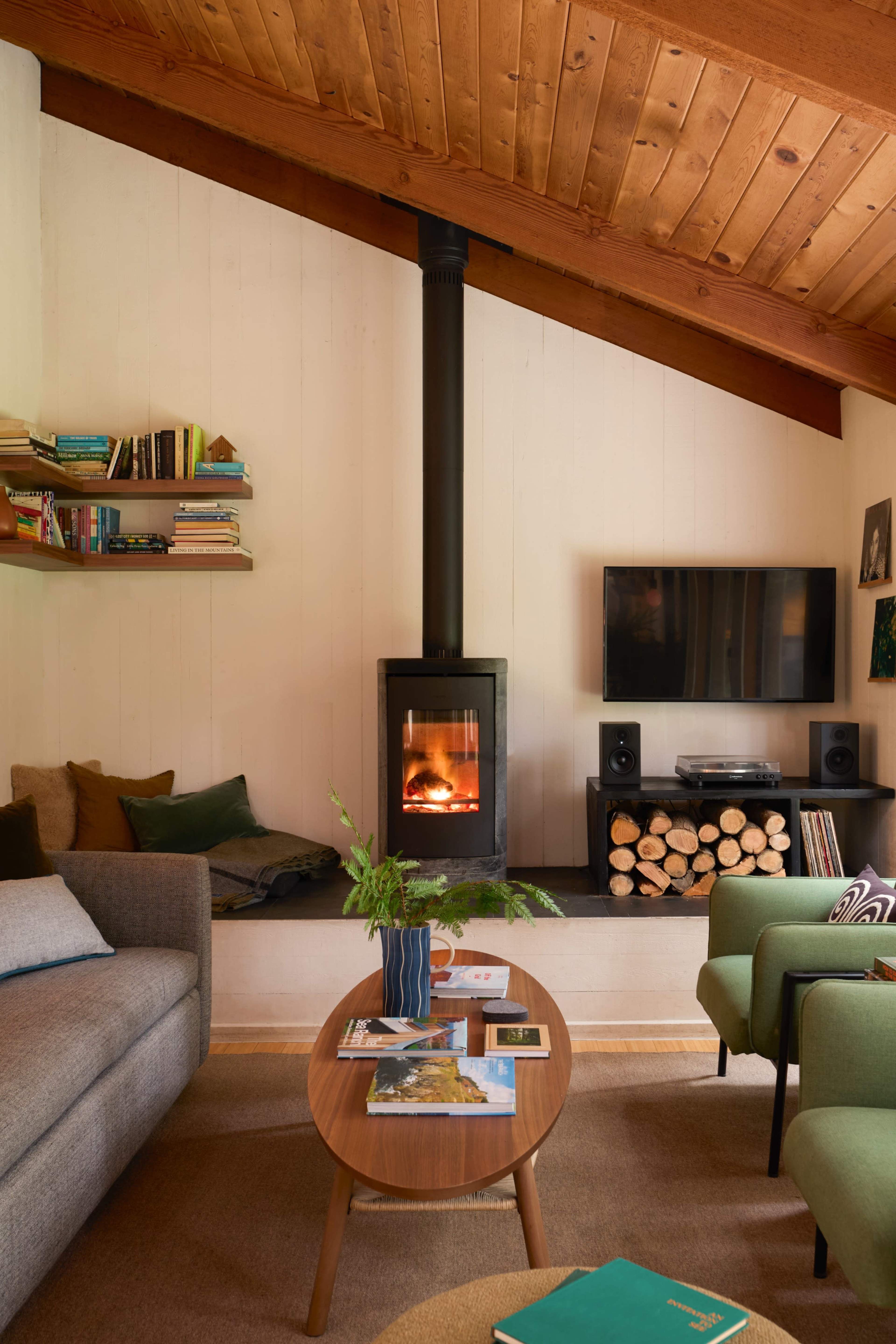 A cozy living room with a modern wood stove, a flat-screen TV, and a table surrounded by green chairs, all set against a backdrop of wooden beams and shelves filled with books.