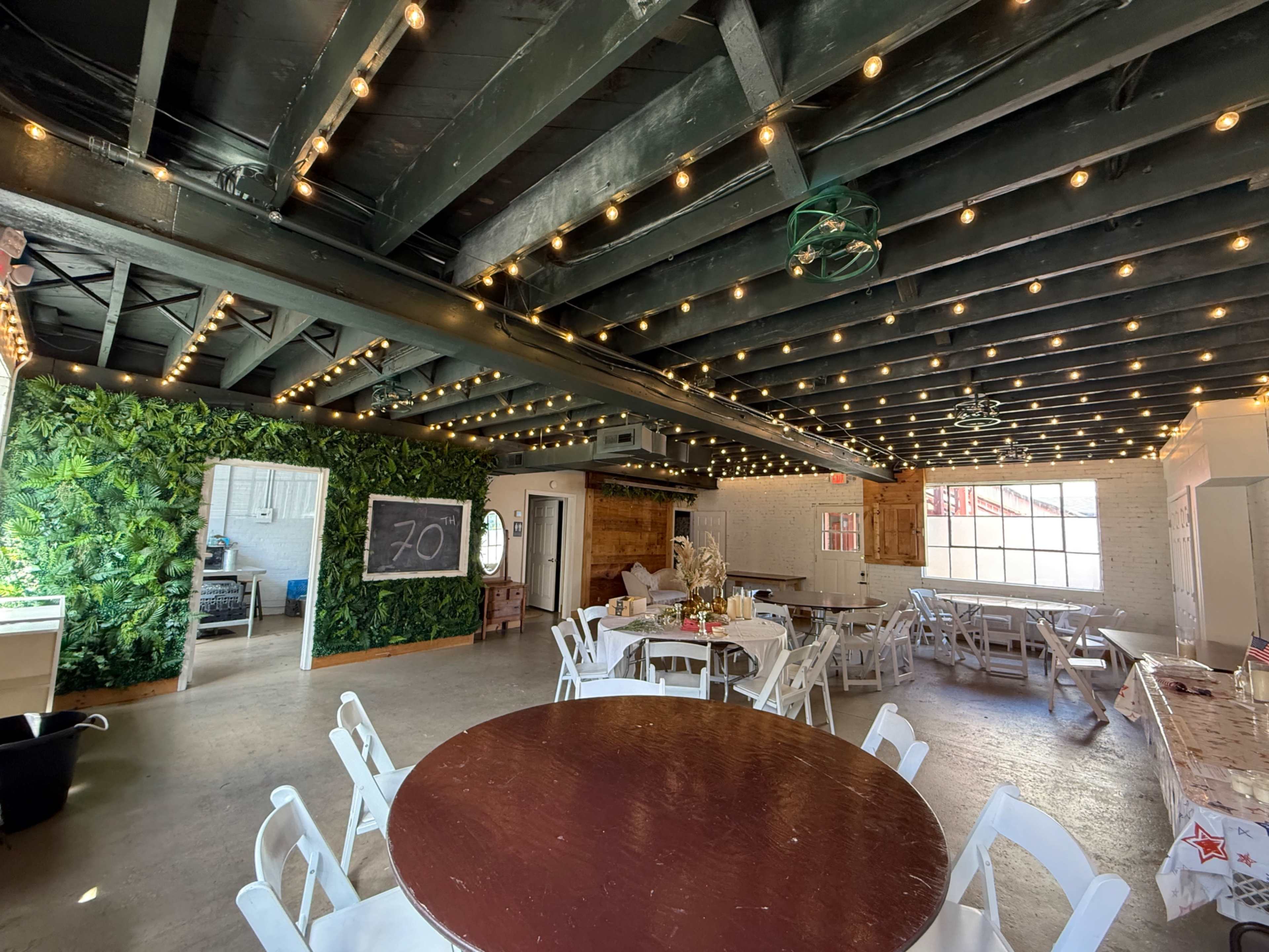 The image shows an indoor event space with string lights overhead, white tables and chairs arranged around dark wooden tables, and a green wall covered in foliage.