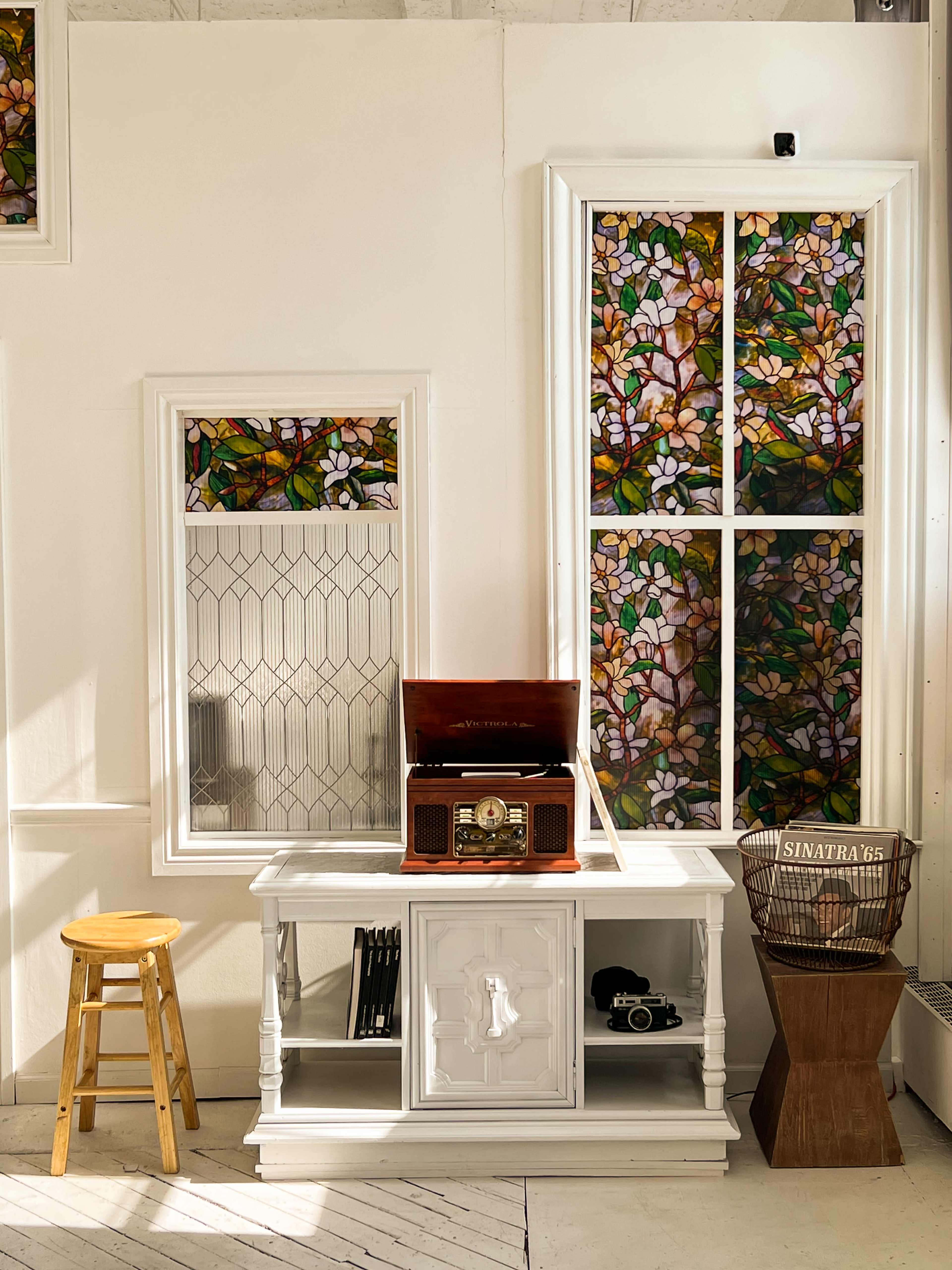 A white room featuring a vintage record player on a cabinet, a wooden stool, and stained glass windows with floral designs.