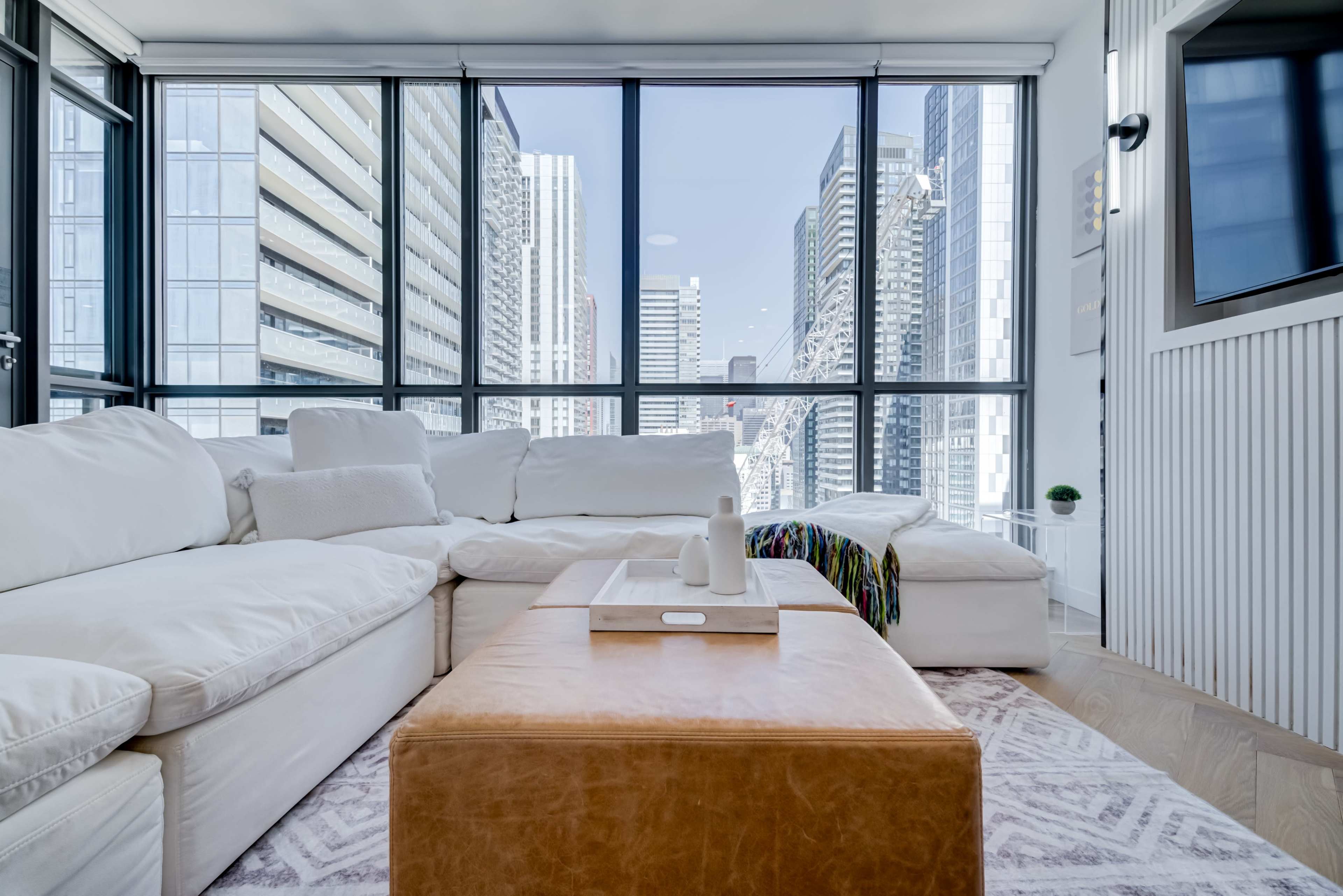 A modern living room features a white sectional sofa, a leather ottoman, and large windows showcasing a city skyline.