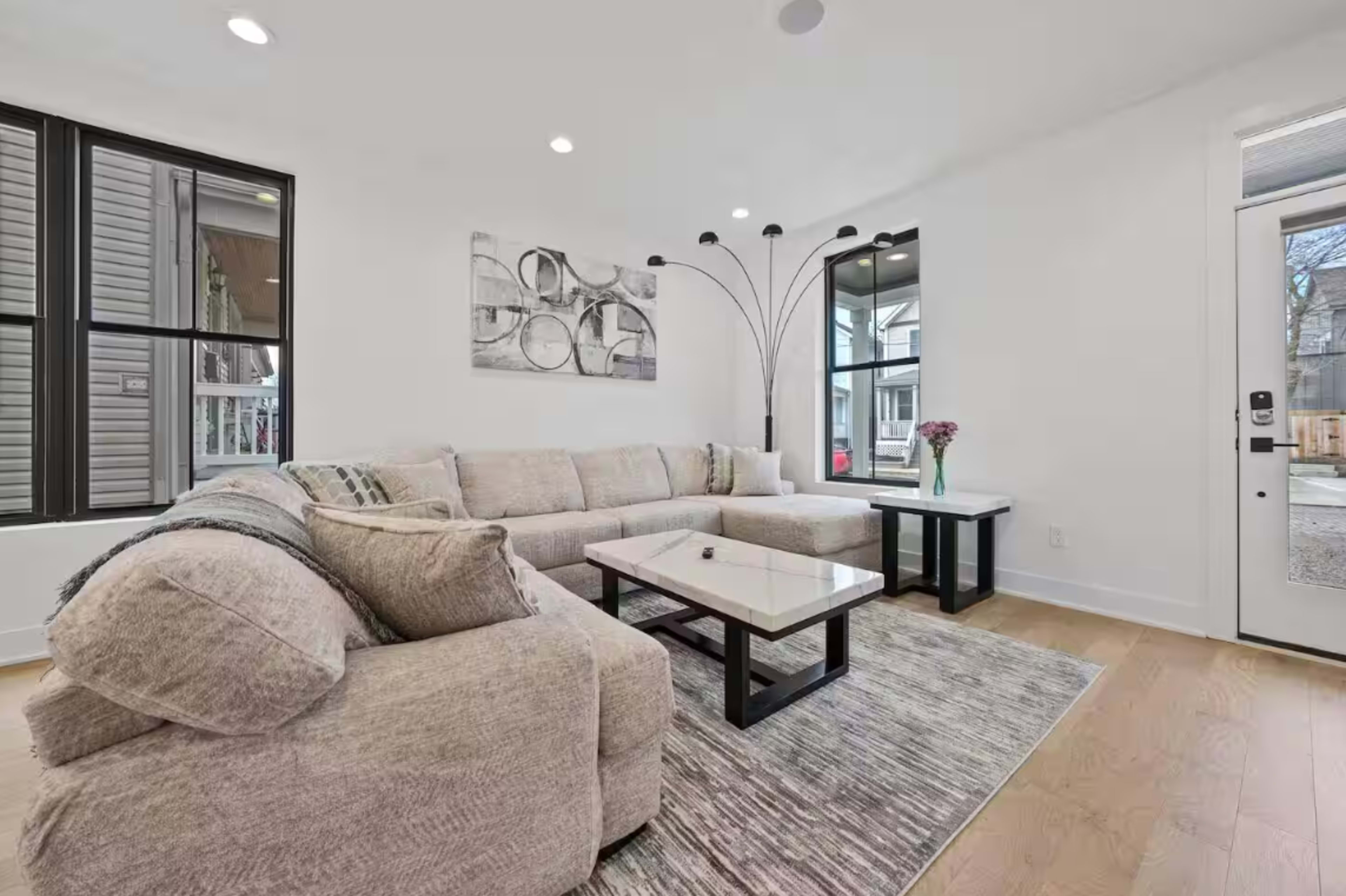 The image shows a modern living room featuring a sectional sofa, a coffee table with a black frame, and large windows allowing natural light to enter.