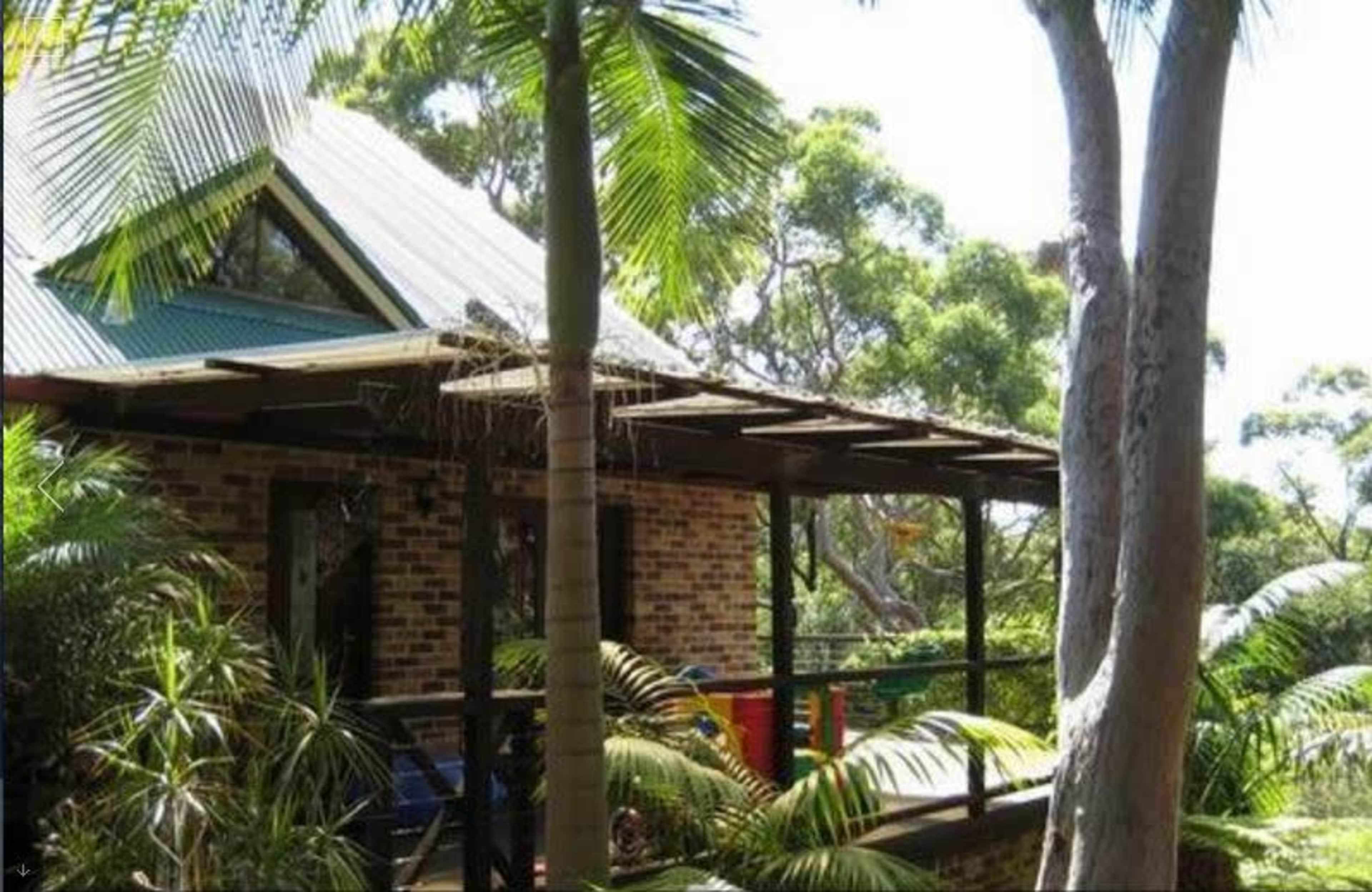 A brick house with a slanted roof is partially surrounded by palm trees and greenery.