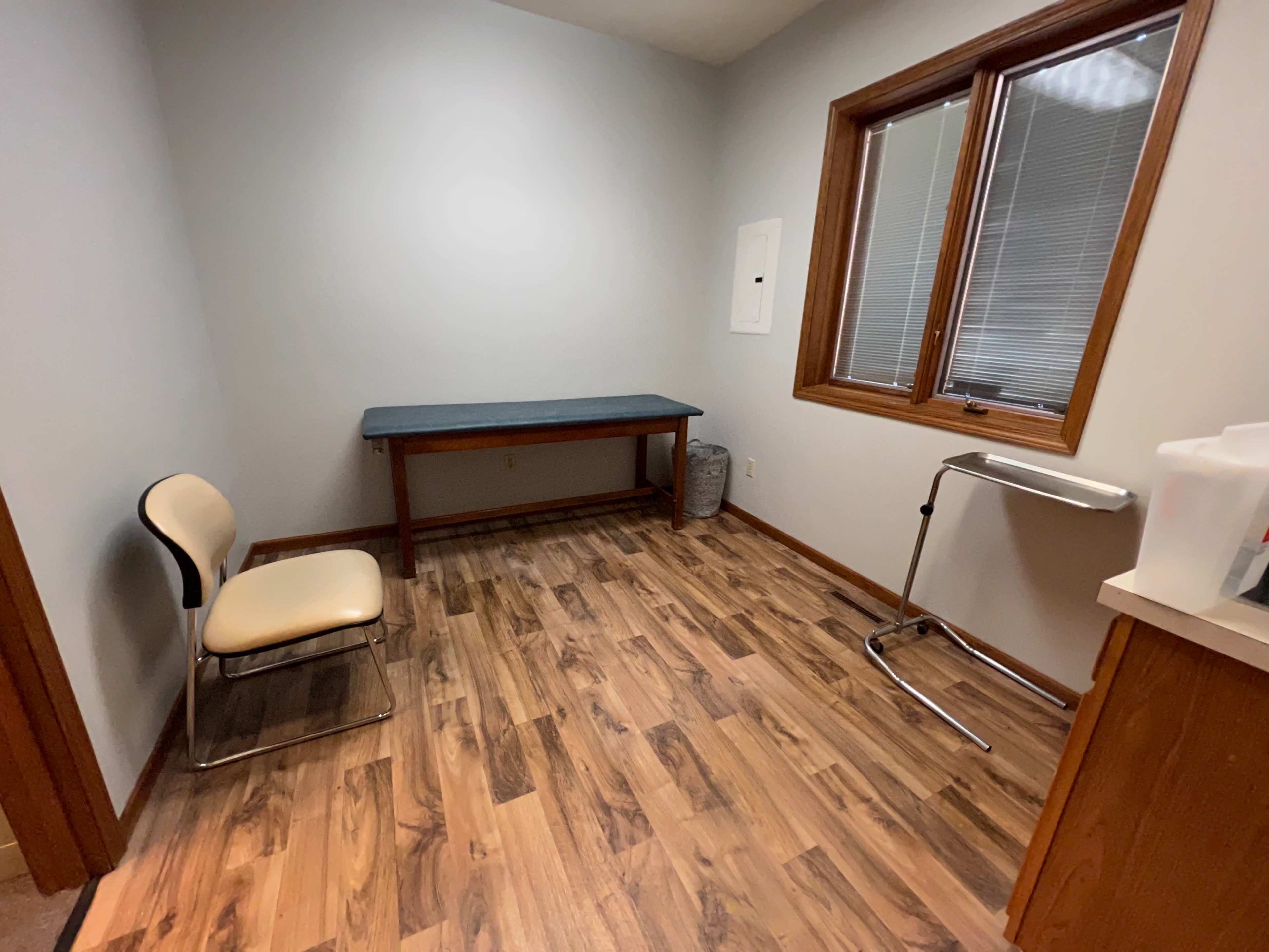 A small examination room with a wooden floor, a table against the wall, a single chair, and a medical stand.