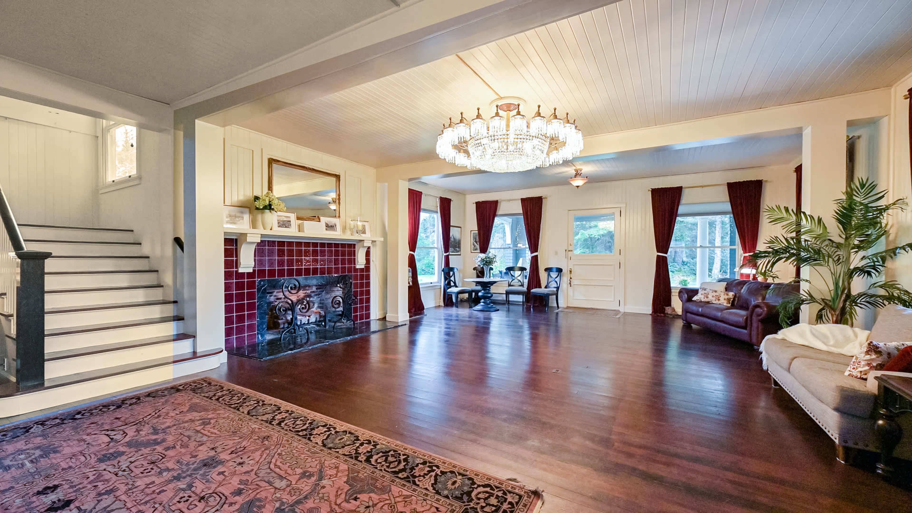The spacious room features a chandelier, a fireplace with decorative tiles, and large windows framed by red curtains.