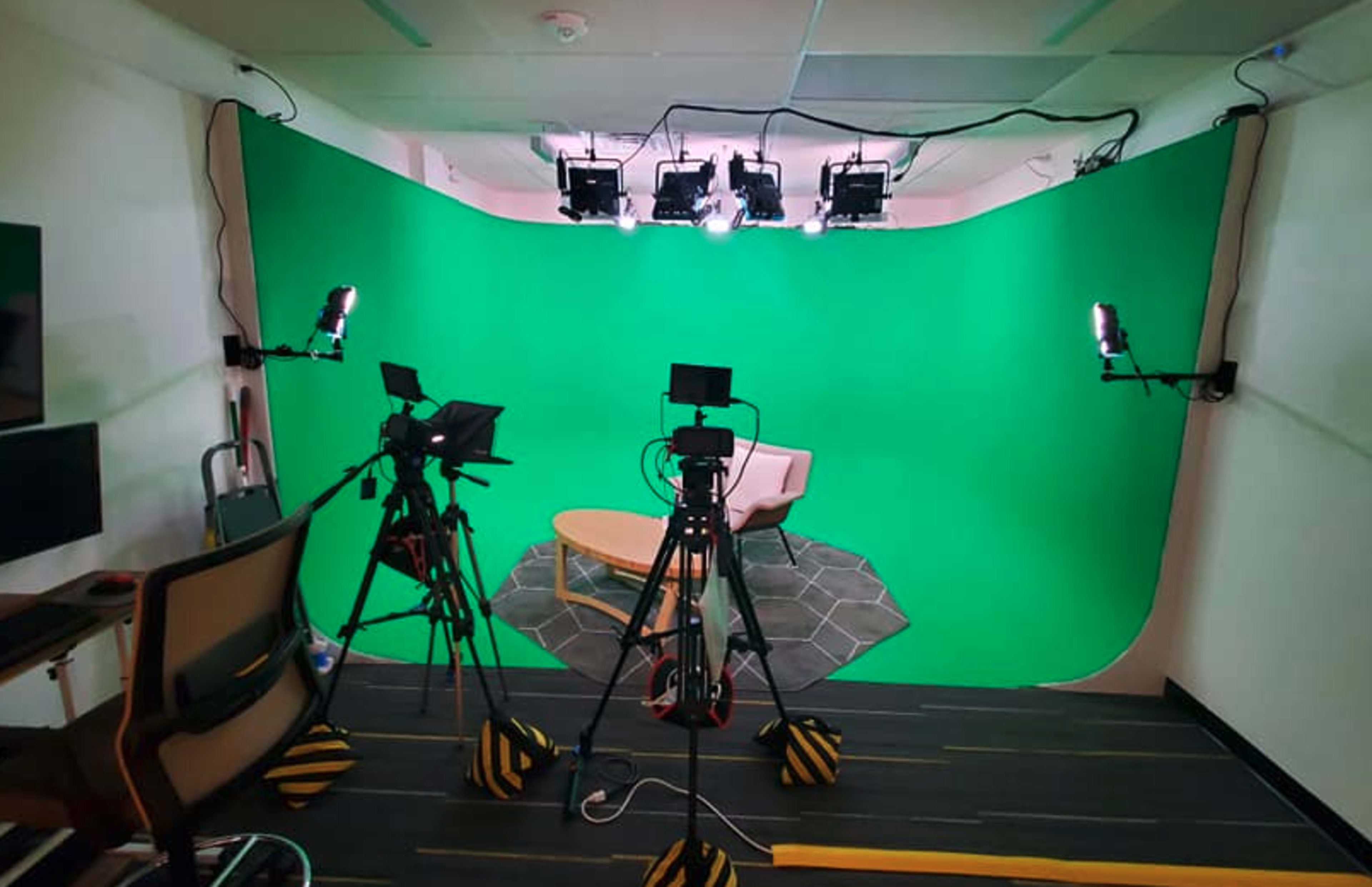 The image shows a video recording studio set up with two tripods holding cameras, a green backdrop, and overhead lights illuminating a small seating area with a beige chair and a coffee table.