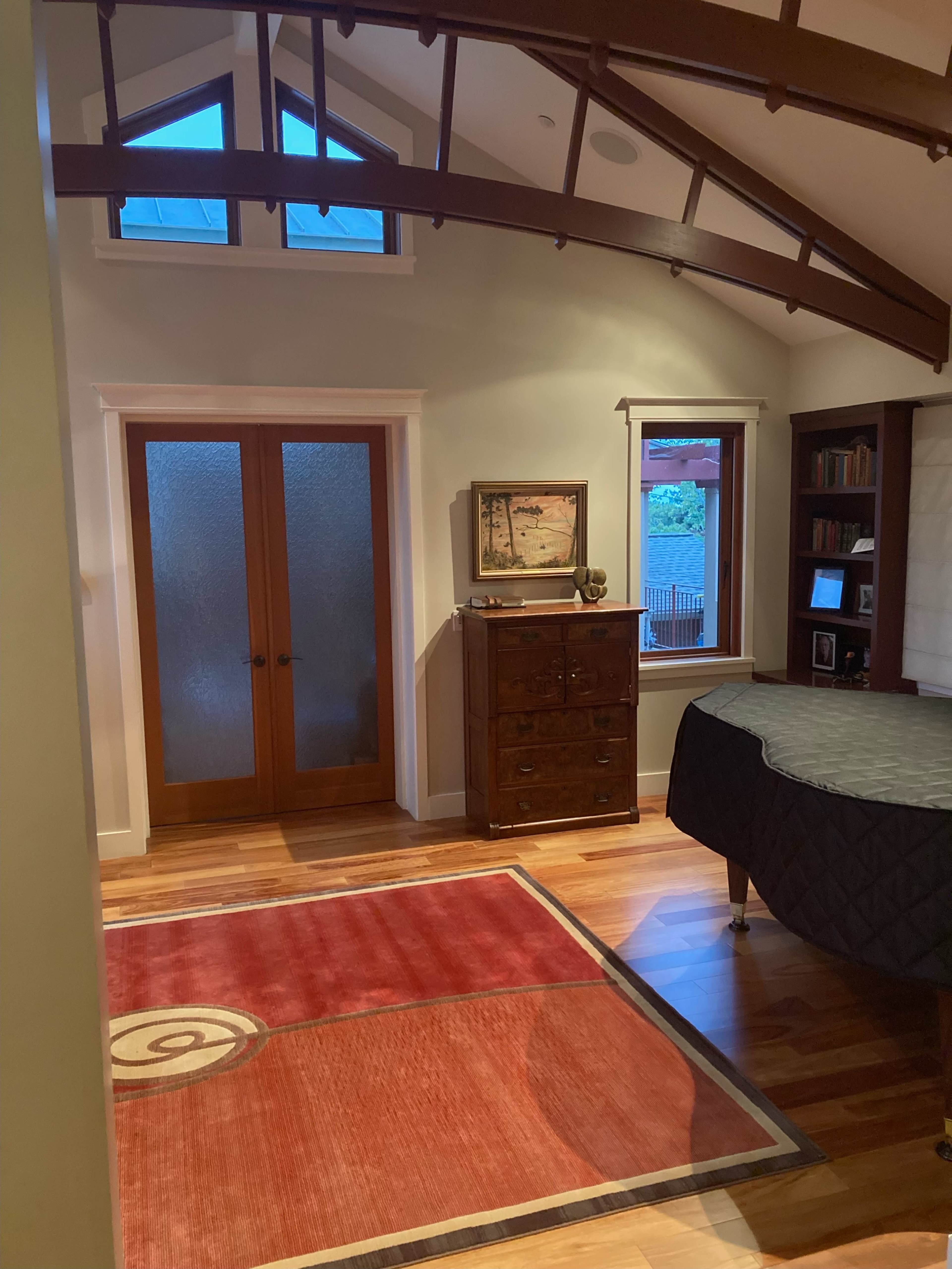 A bedroom with wooden flooring, sloped ceilings, a large rug, and a set of double doors leading to an outdoor area.