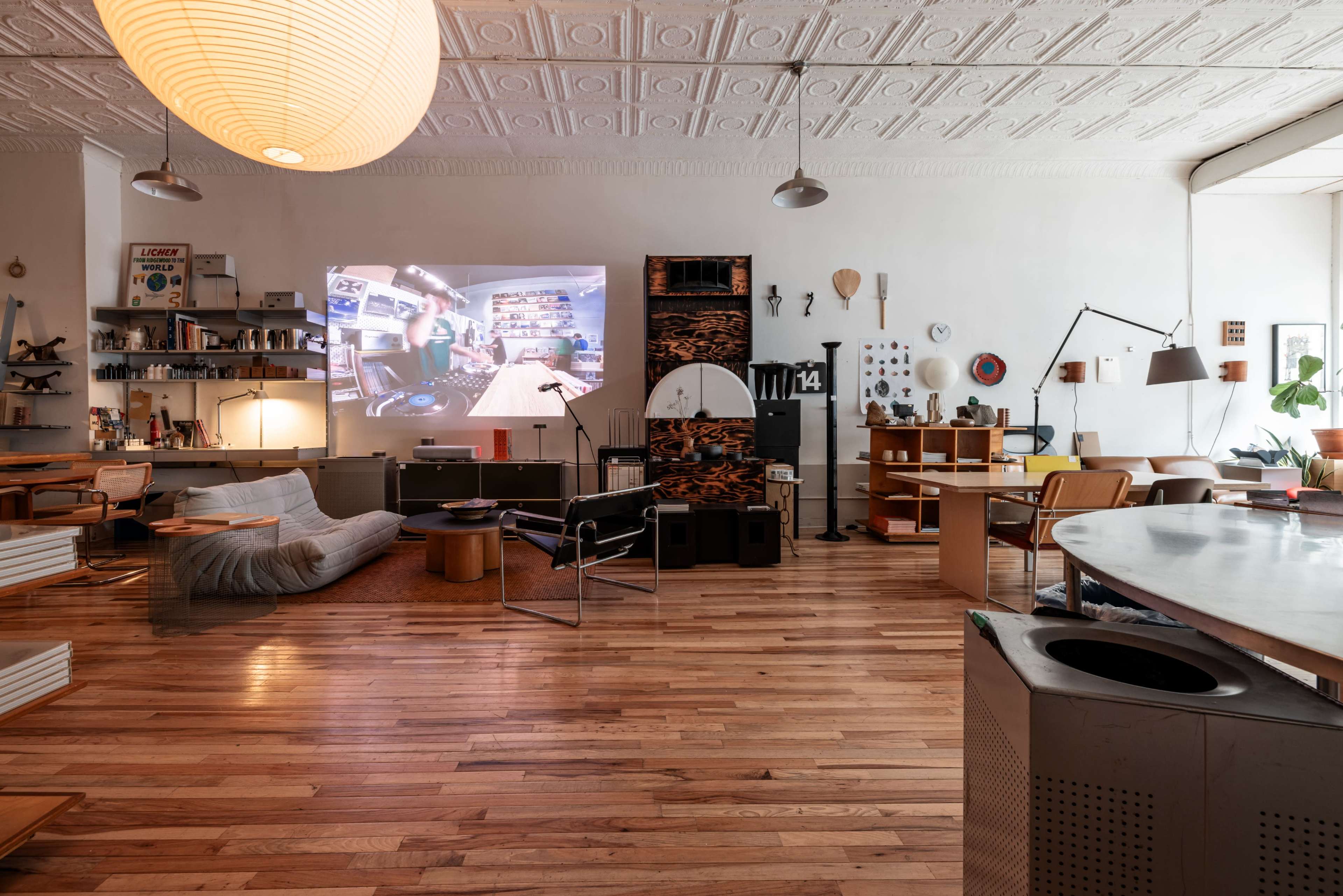 A spacious, modern living area with wooden floors, featuring a large projection on the wall, various furniture pieces, and shelves filled with books and decor.