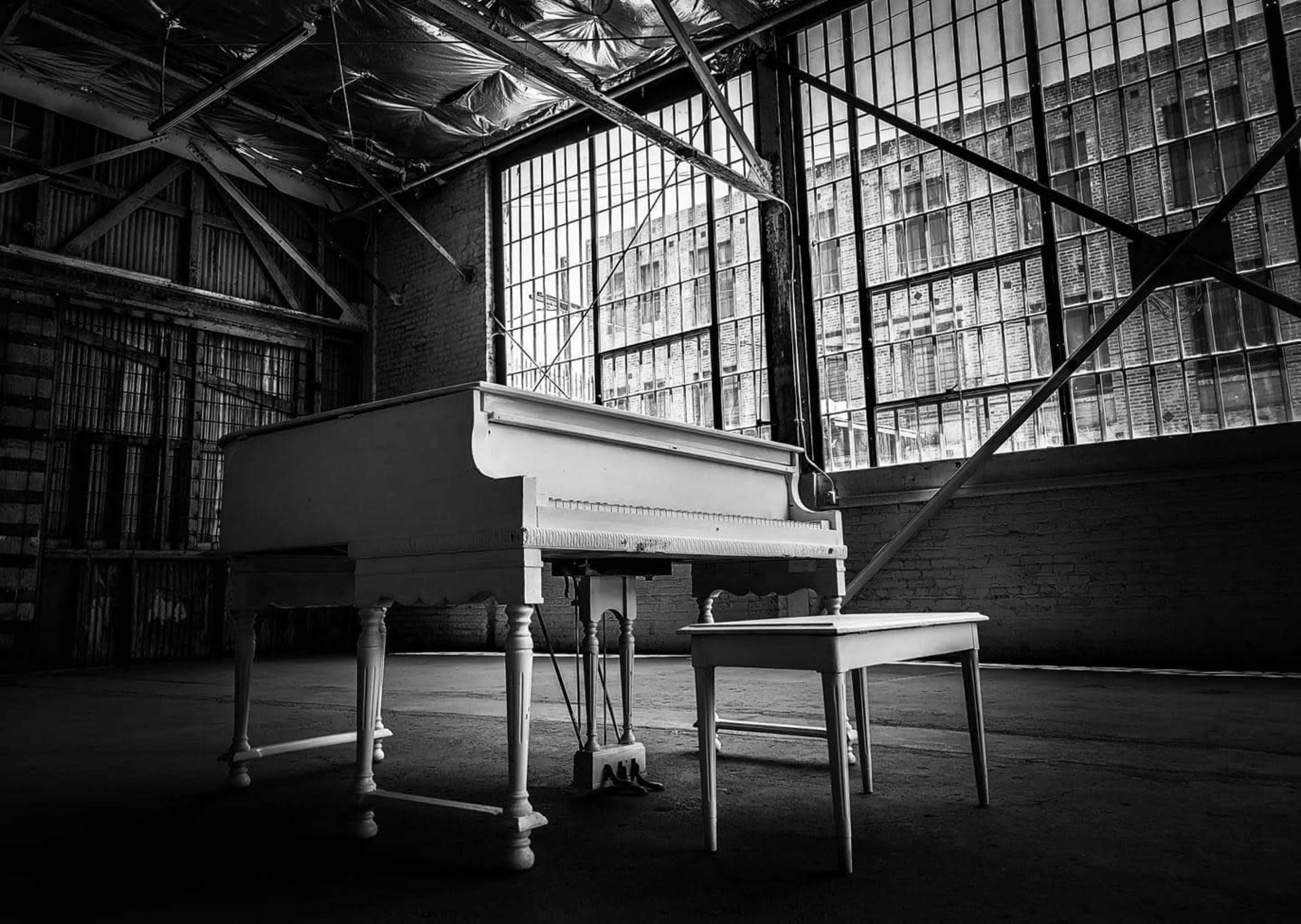The image shows a white grand piano and a matching bench placed in an empty warehouse with large windows.