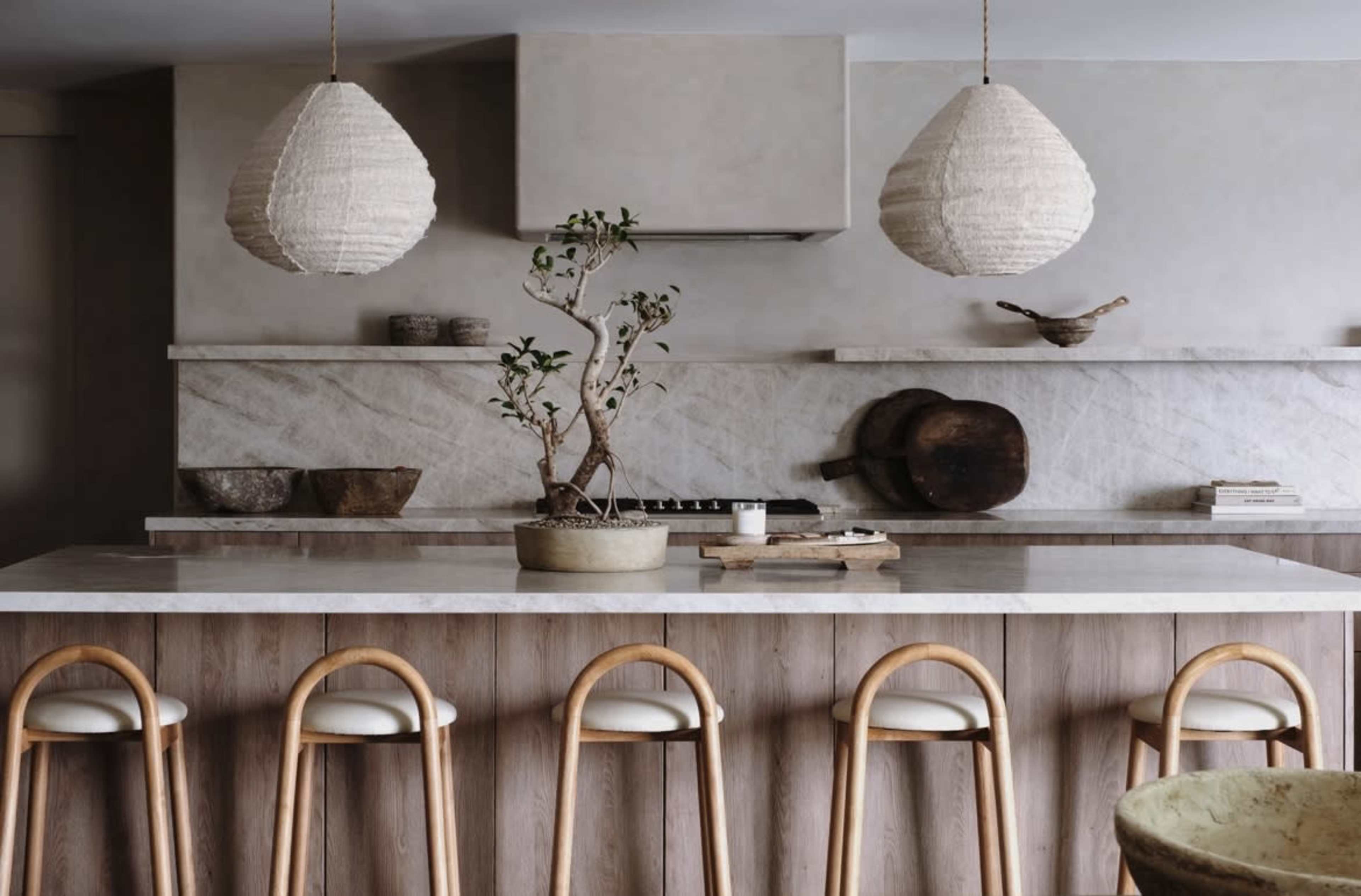 The image shows a modern kitchen with a large marble island, wooden bar stools, and pendant lighting, featuring a minimalist design with natural elements.