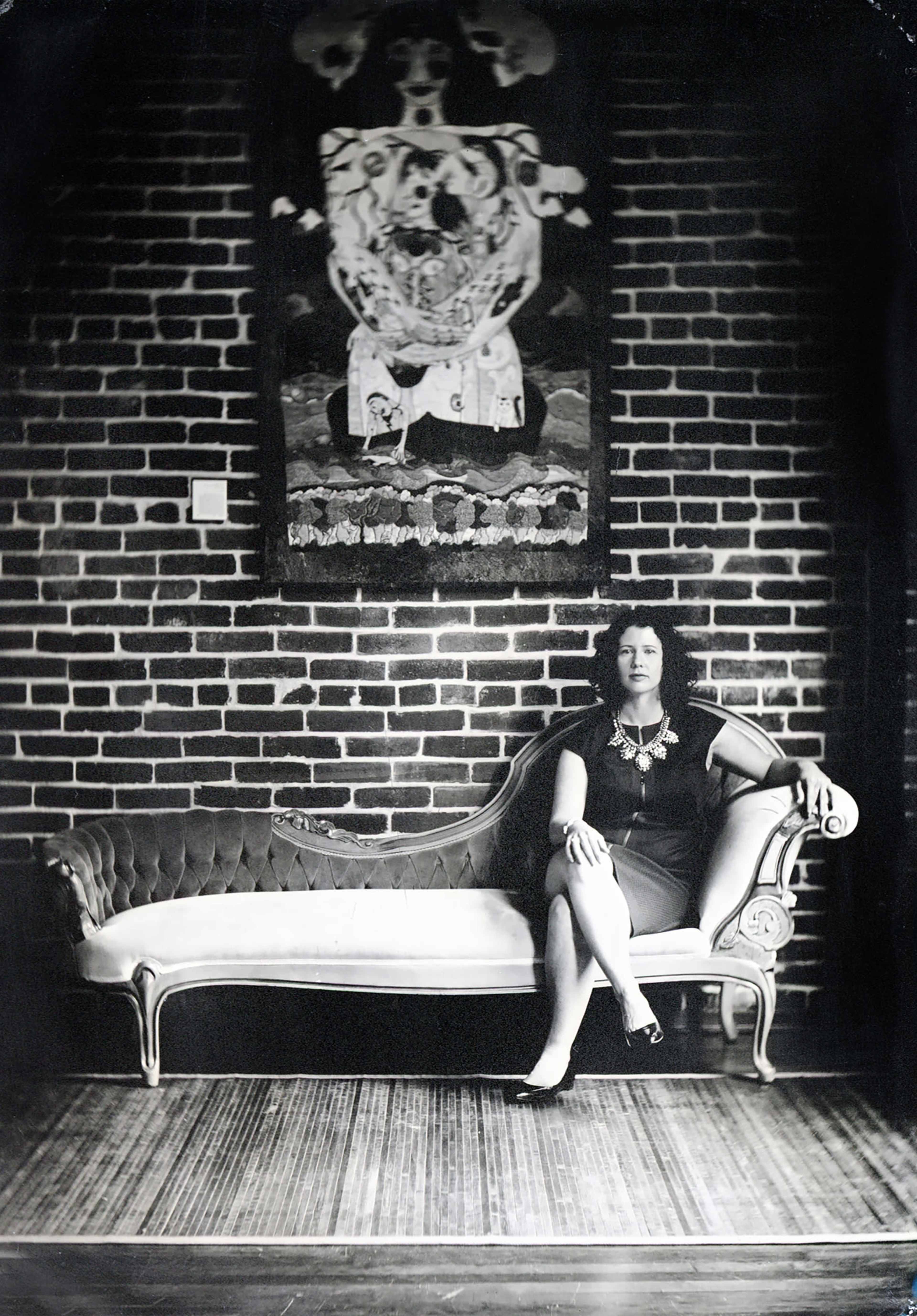 A woman sits on an elegant, vintage sofa in front of a brick wall adorned with a large artwork.