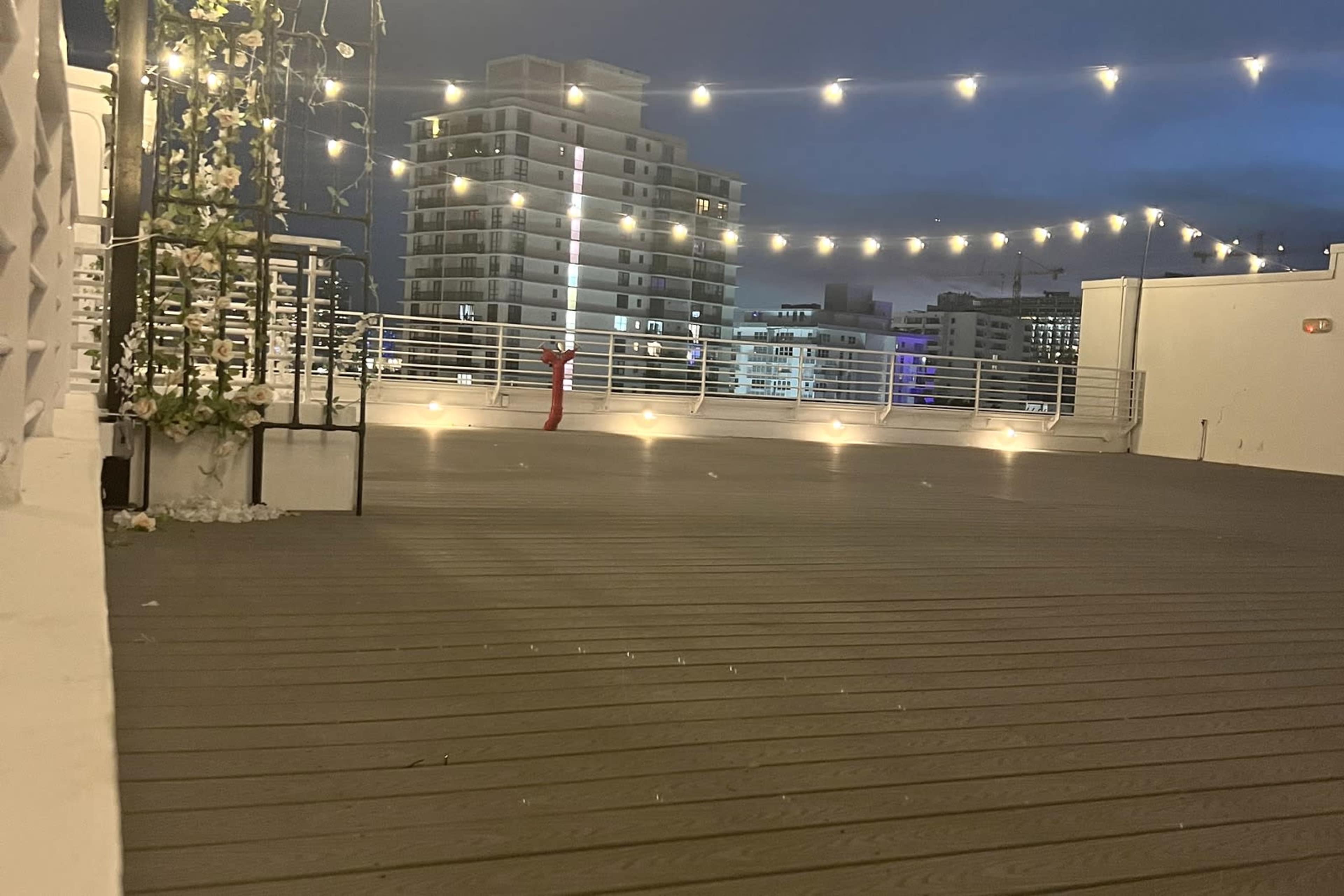 The image shows a rooftop area with wooden flooring, illuminated by string lights, overlooking city buildings at night.