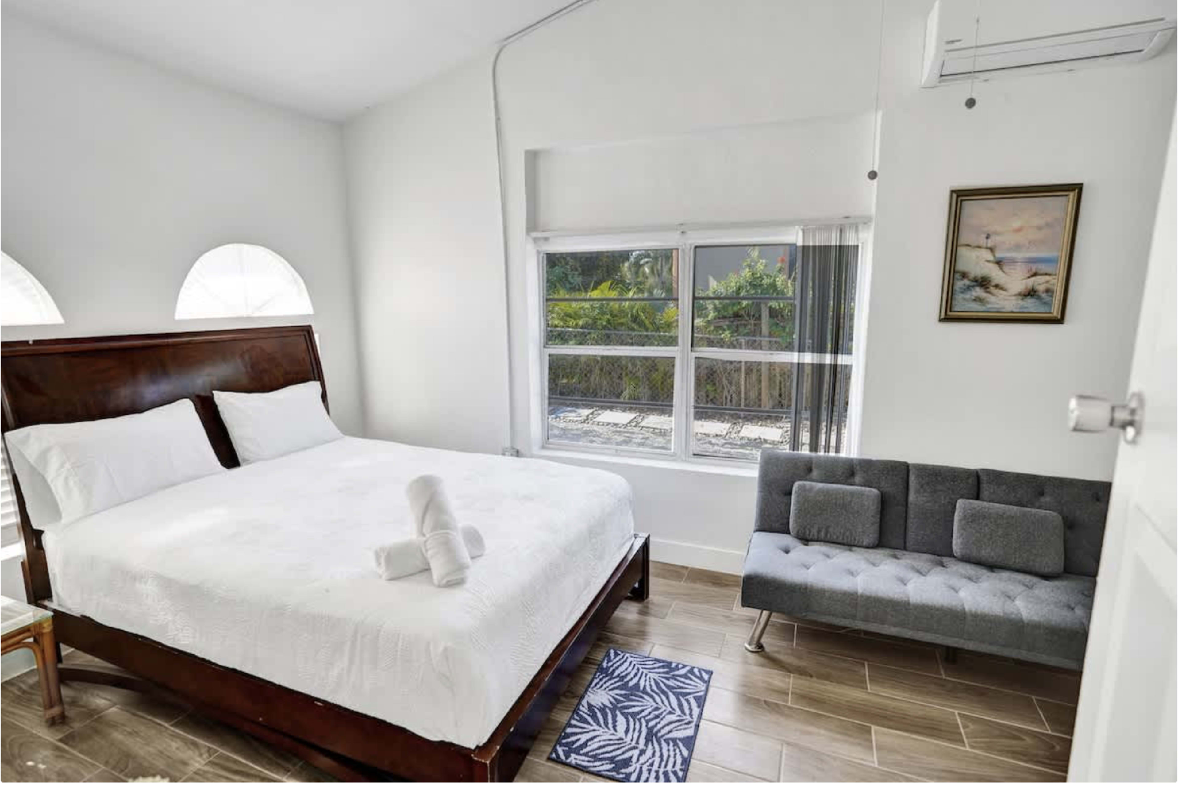 The image shows a simple bedroom featuring a white bed with rolled towels, a gray sofa, and a window with natural light.