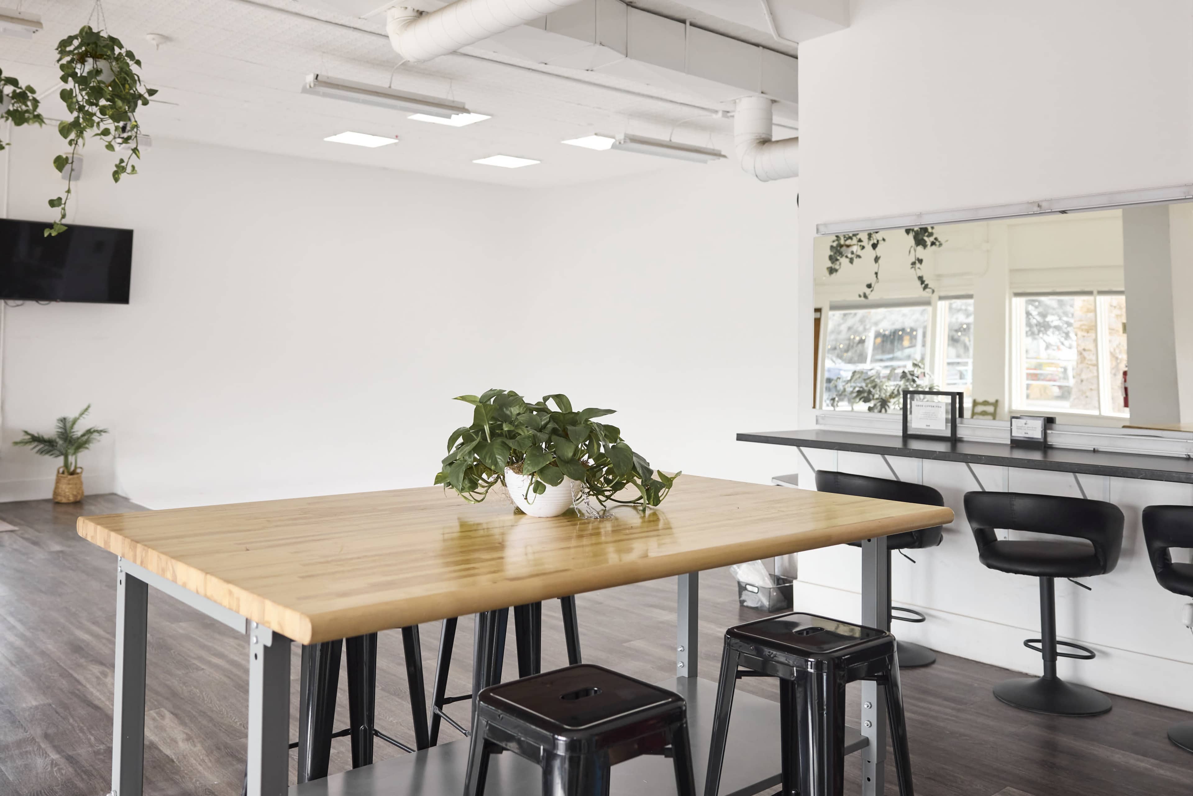 A wooden table with black stools and a potted plant is positioned in a bright, minimalist room featuring white walls and a large mirror.