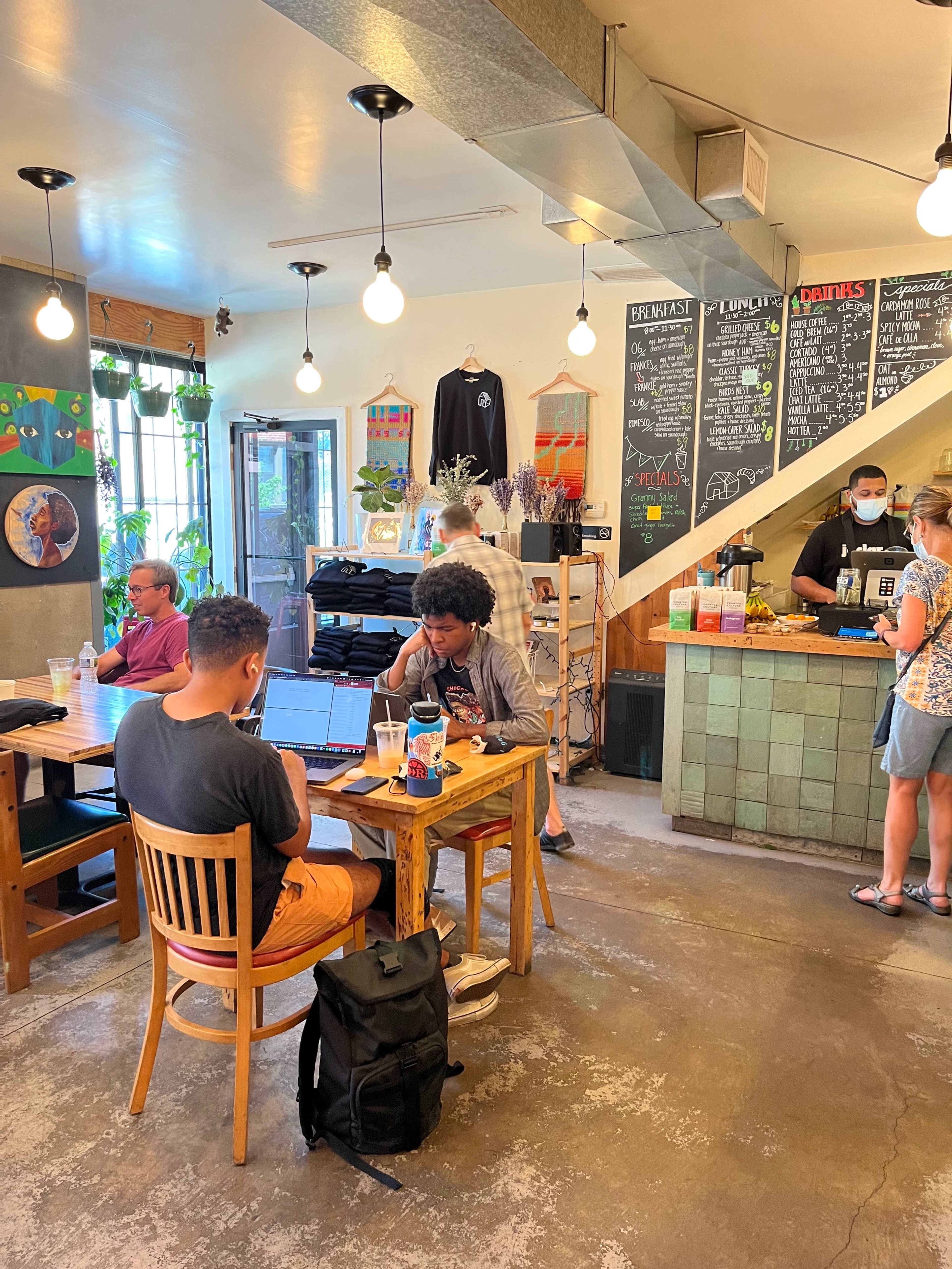 A bustling café features patrons working at tables, a barista serving customers at the counter, and a menu board on the wall.
