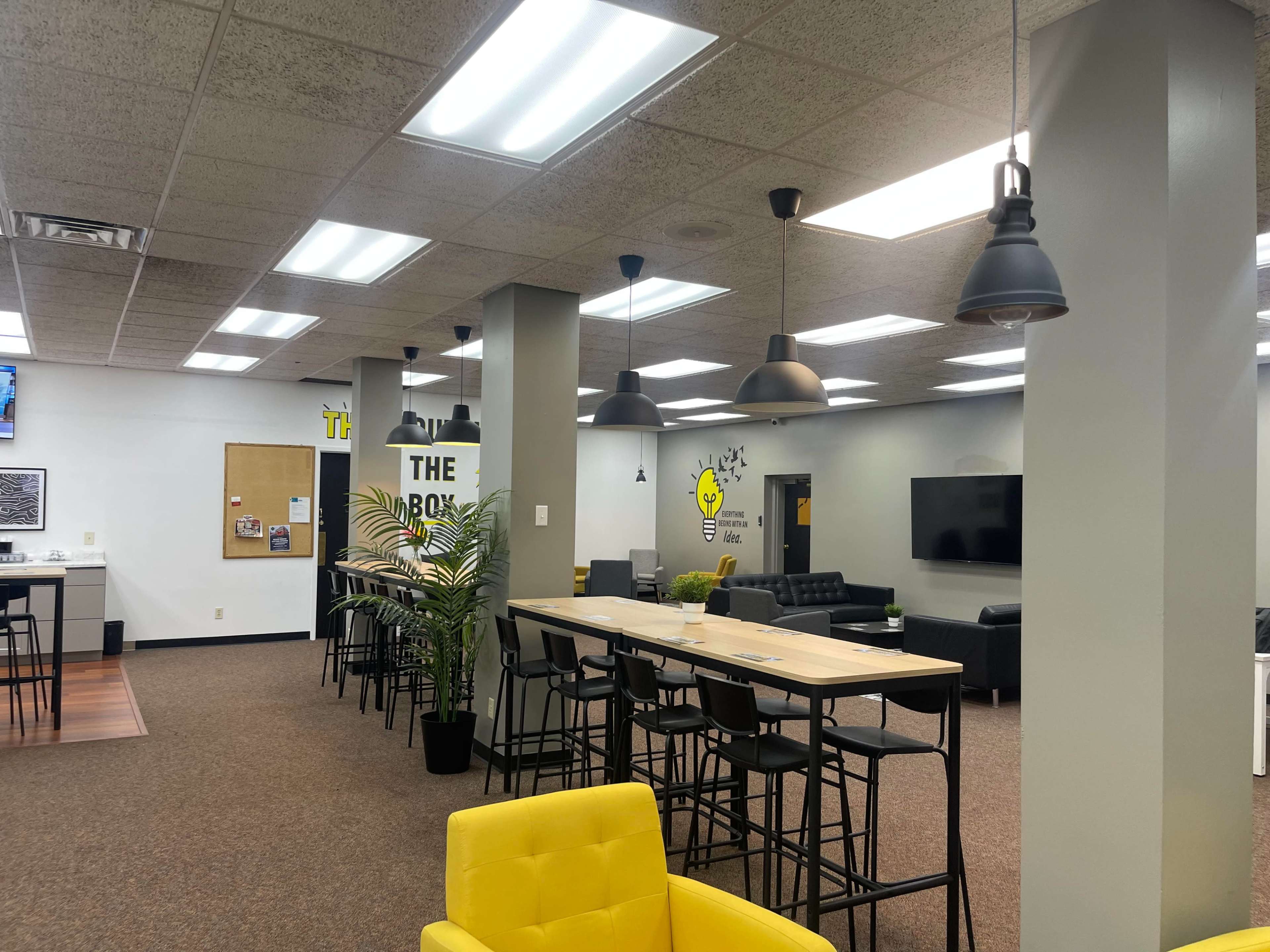 The image shows an open, modern workspace featuring a kitchen area with bar stools, a communal table, and seating areas with sofas.