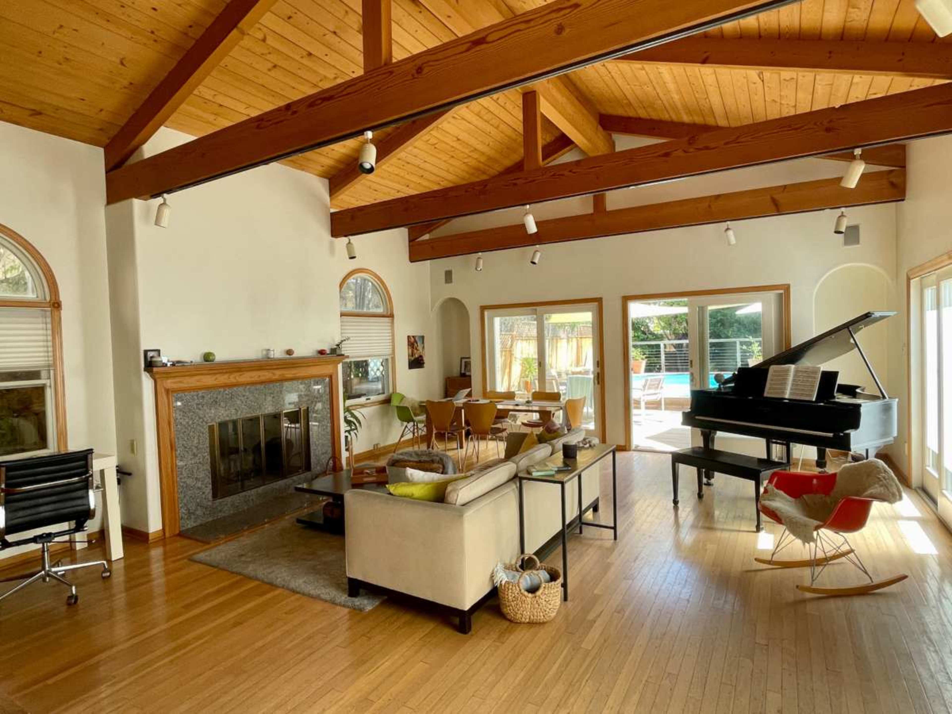 The spacious living room features a grand piano, a fireplace, and large windows that overlook an outdoor pool area.