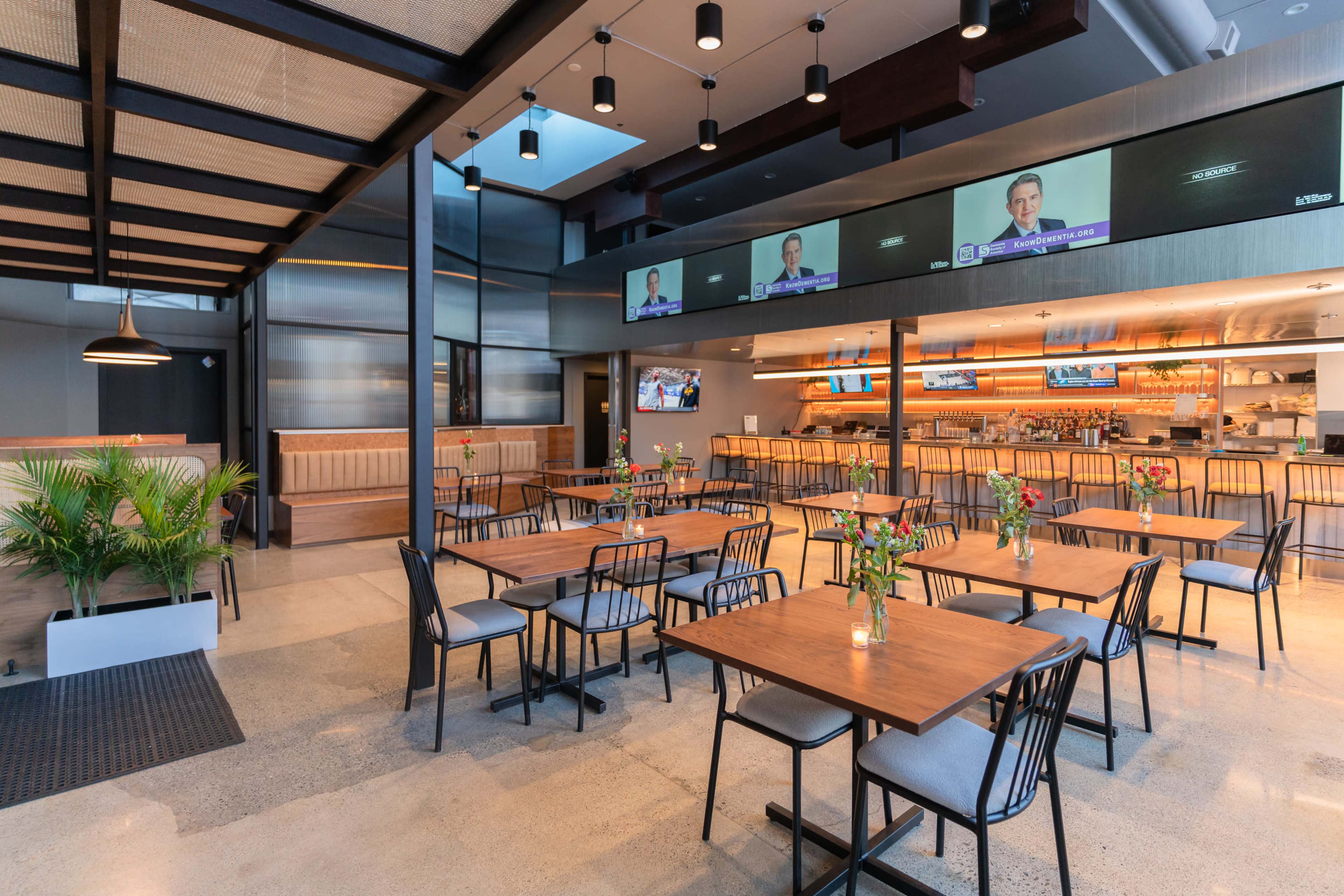 The interior of a modern restaurant features several empty wooden tables and chairs, a large bar area in the background, and multiple screens displaying news.