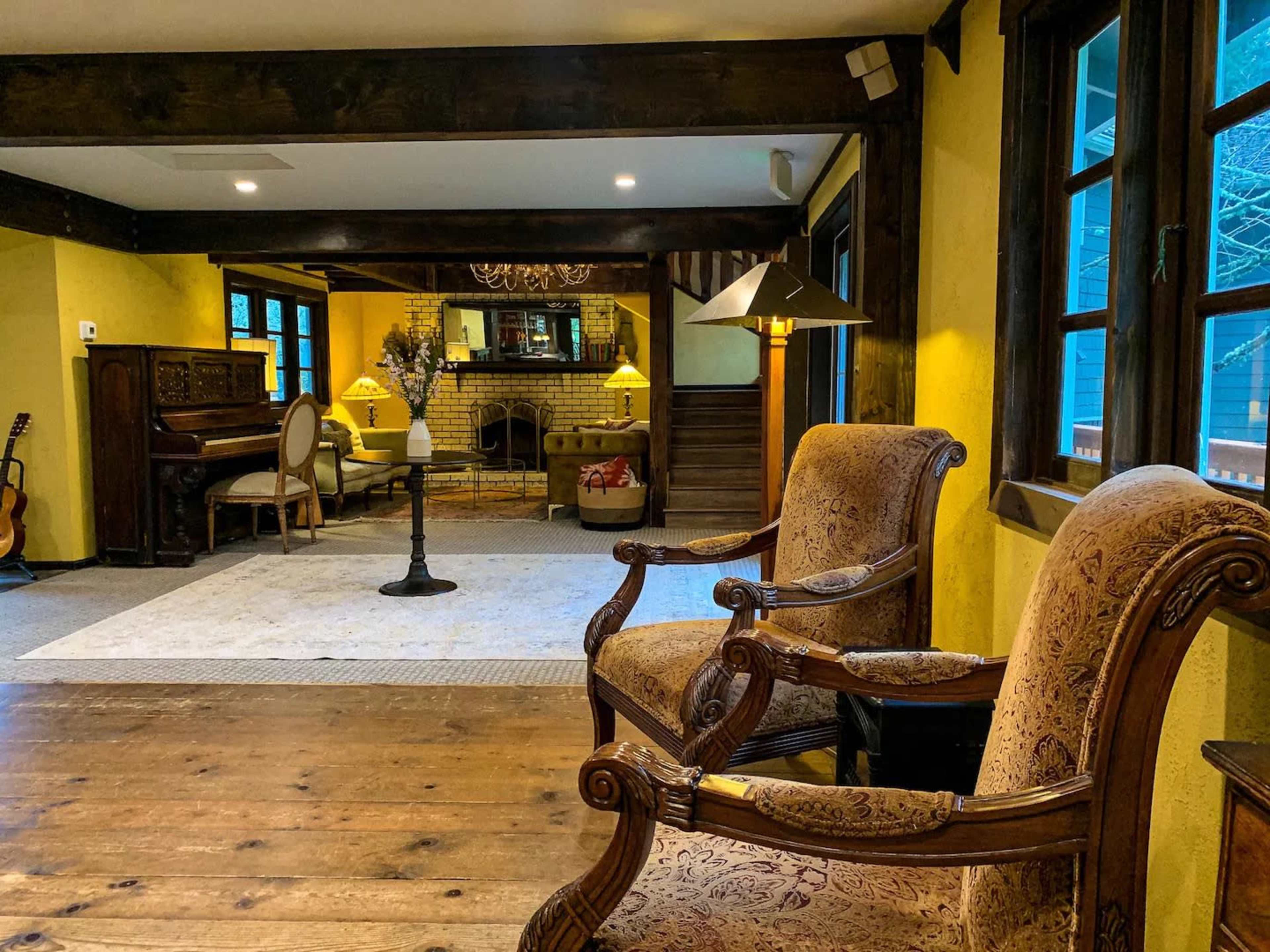 The image shows a cozy living room with wooden beams, two upholstered chairs, a fireplace, a piano, and large windows.