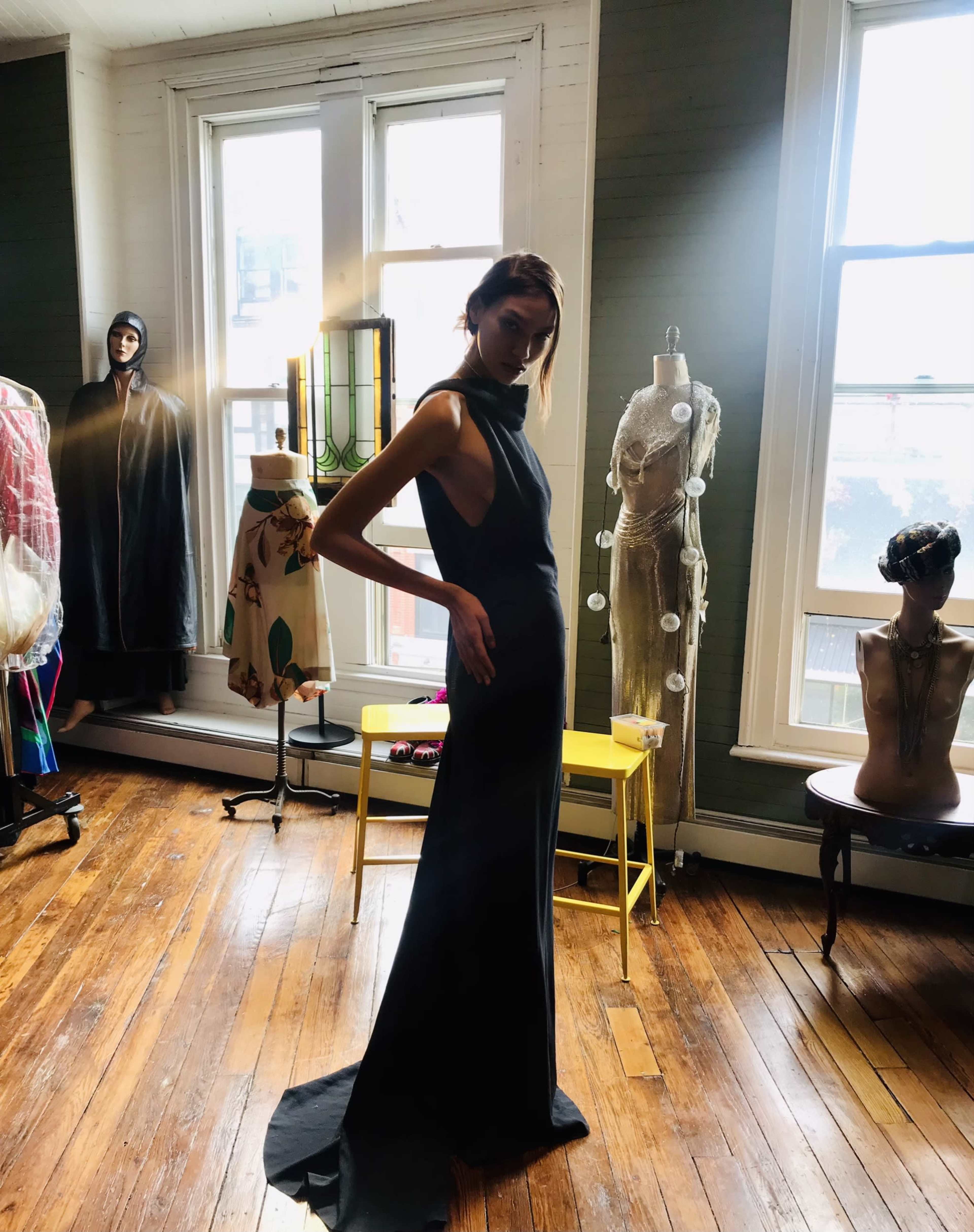 A model poses elegantly in a black gown inside a well-lit room filled with various dress forms and clothing.