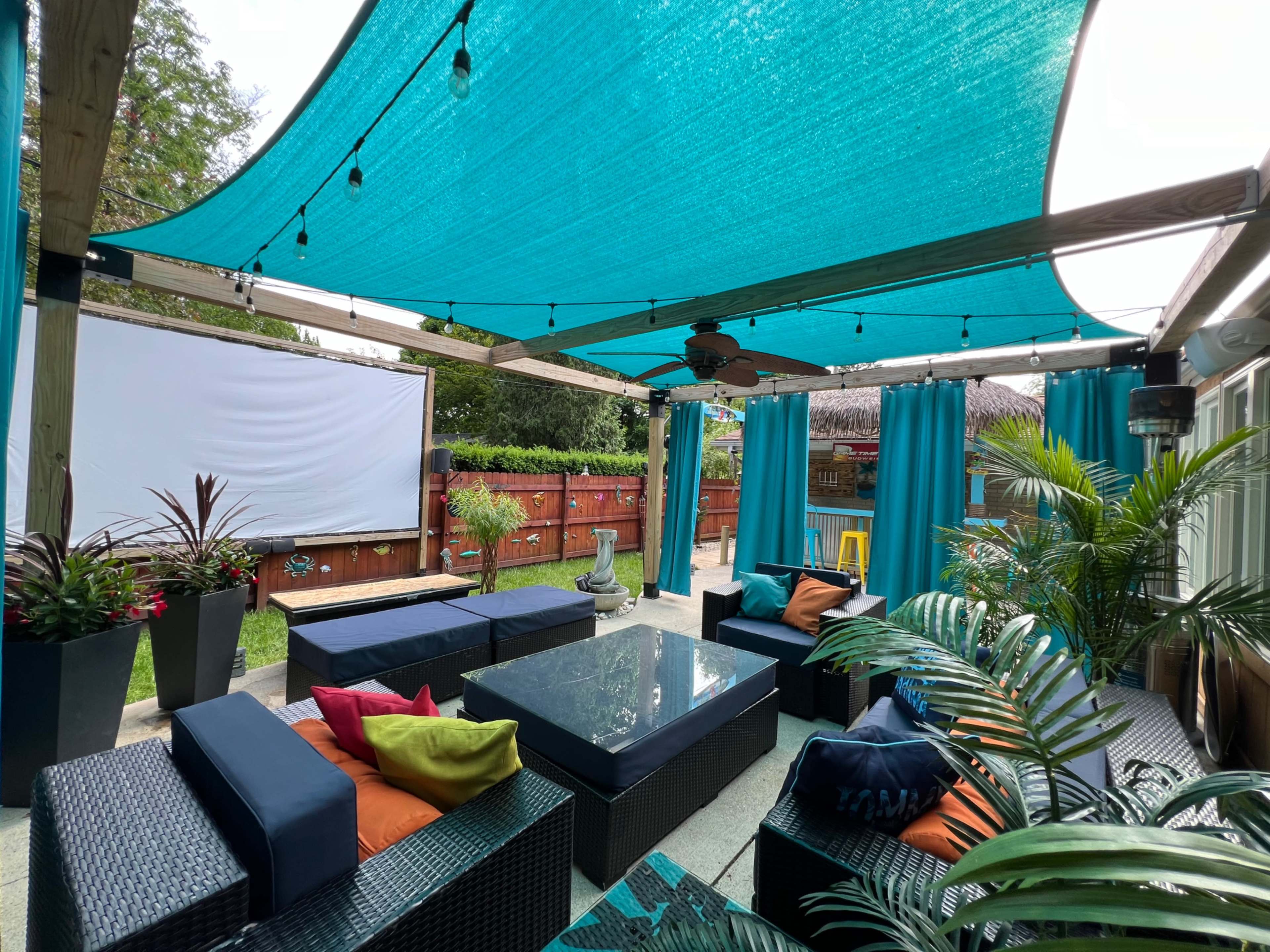The image shows a patio area with blue shade sails overhead, a large outdoor screen, and dark wicker furniture arranged around a table surrounded by plants.