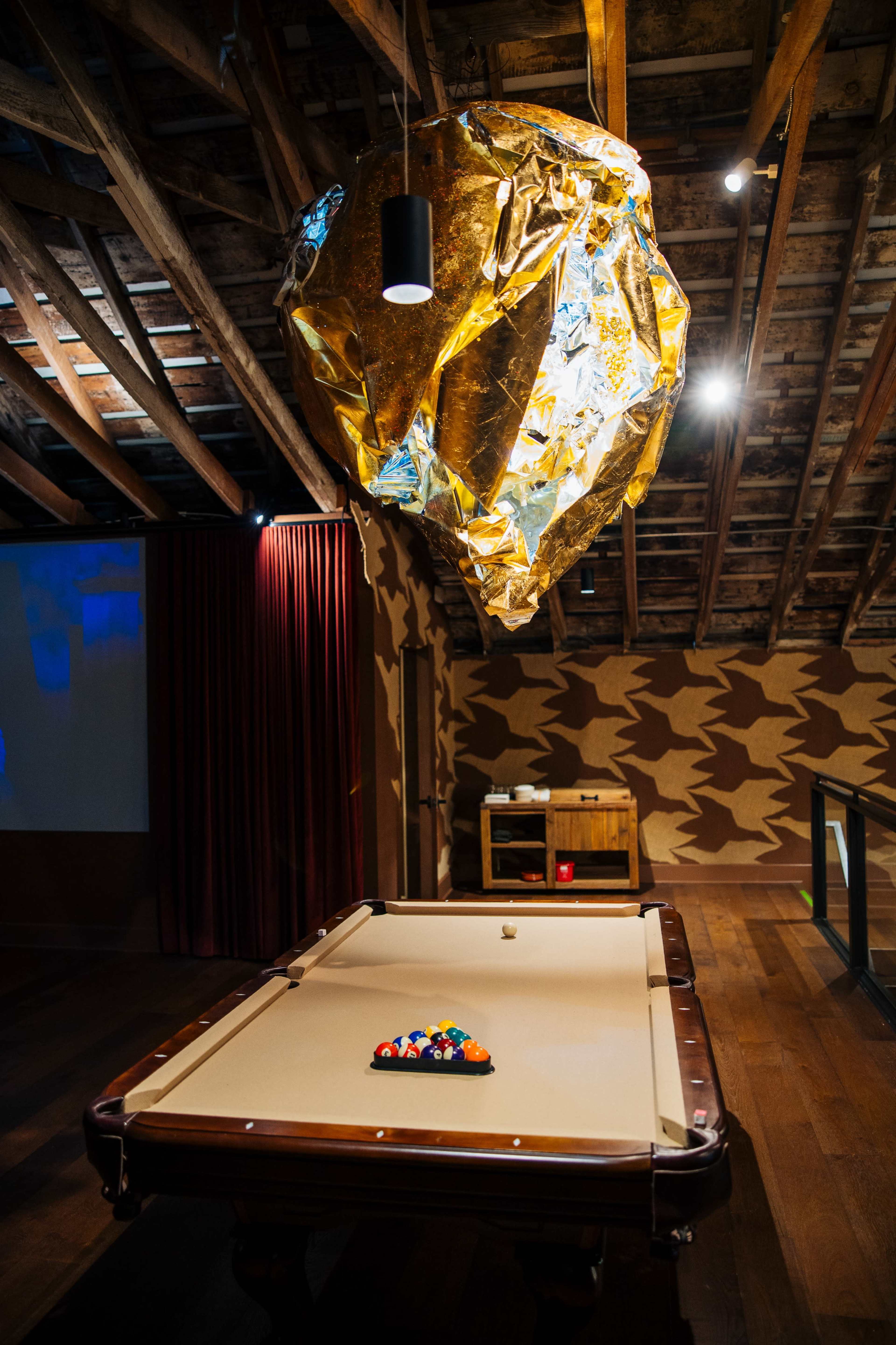 The image shows a billiard table with a rack of balls arranged on it, beneath a metallic gold sculpture hanging from the ceiling in a rustic room with exposed wooden beams.