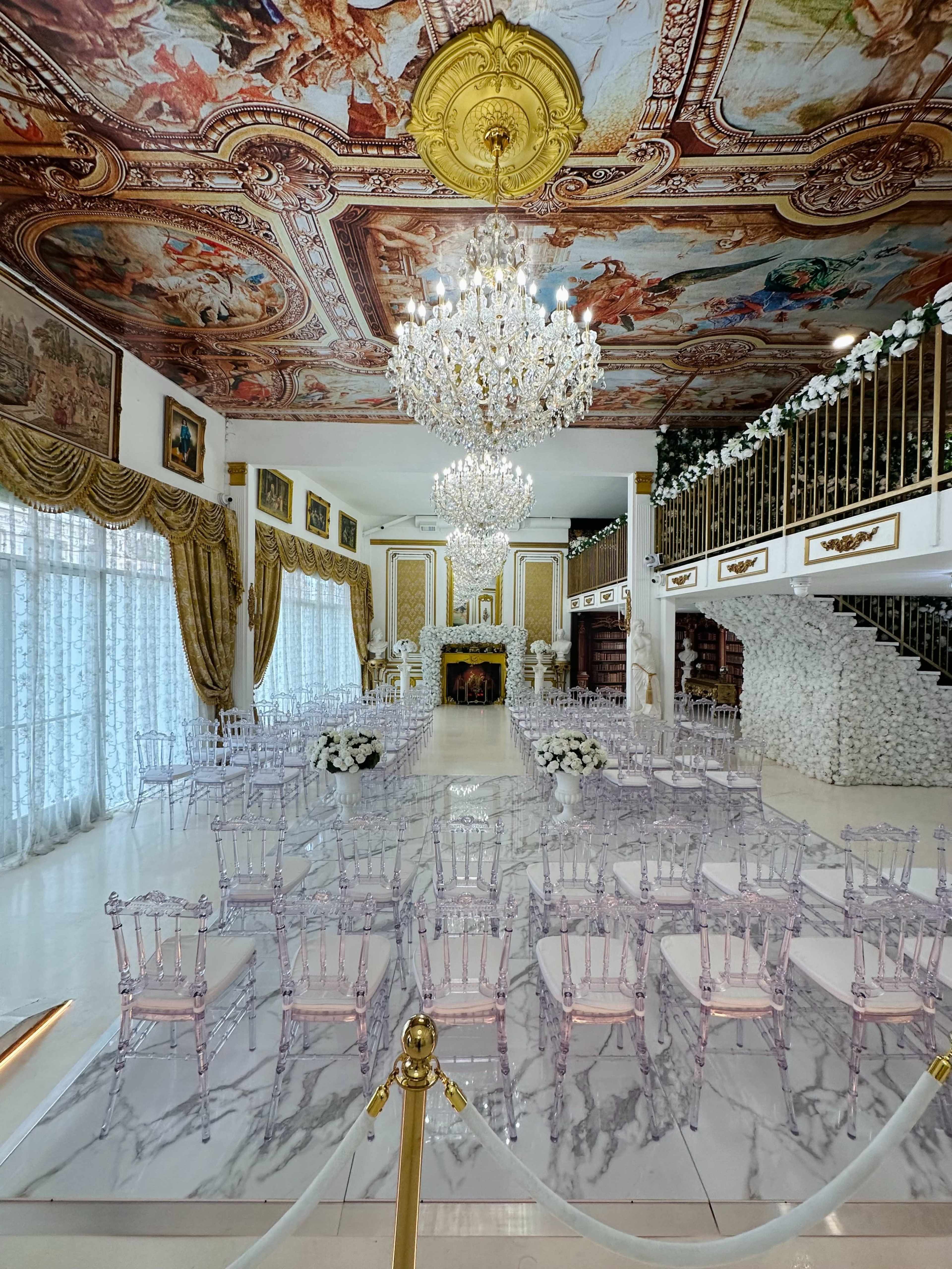 The image shows an ornate indoor venue with crystal chandeliers, a painted ceiling, and rows of clear chairs arranged for a ceremony.