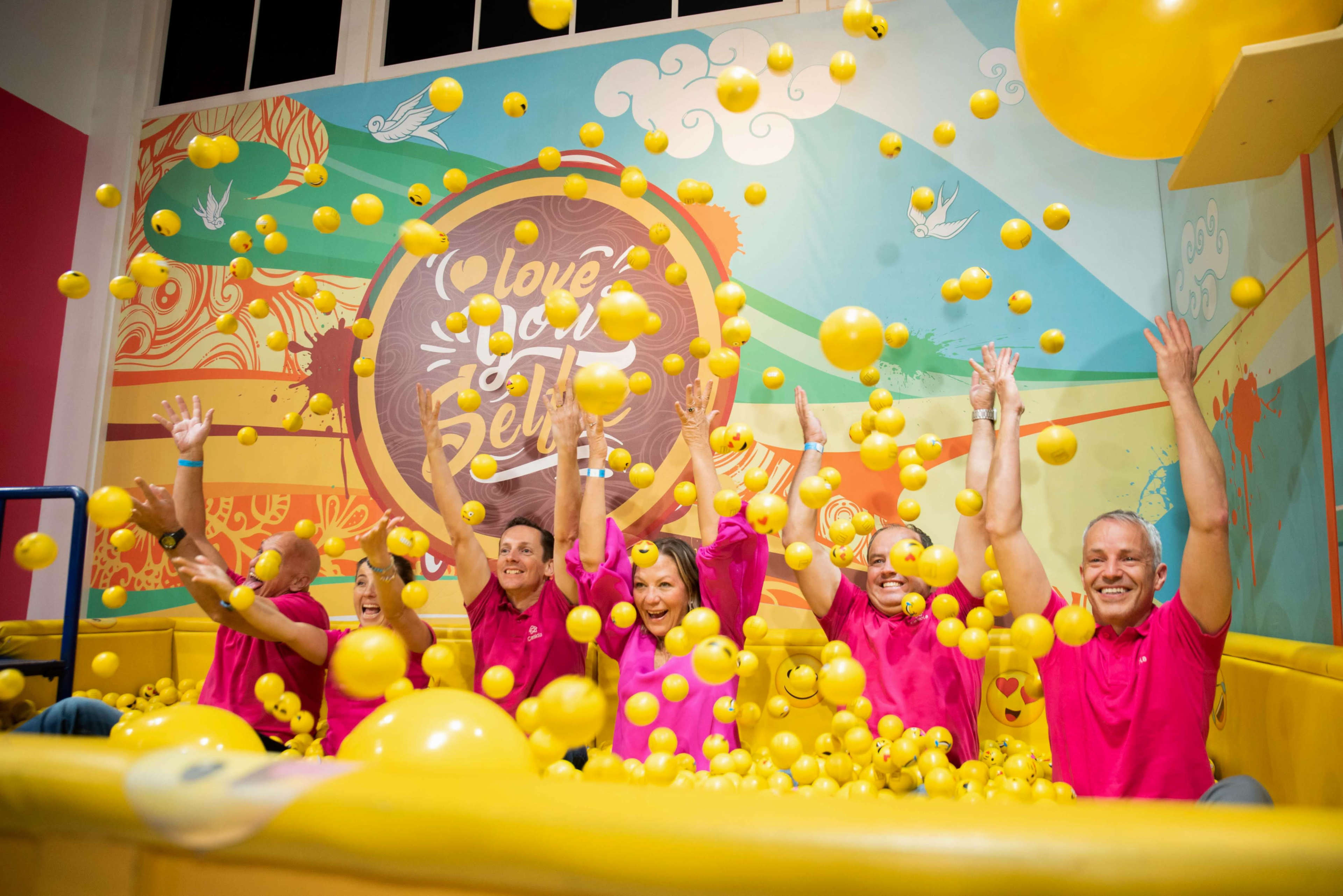 A group of six people in pink shirts joyfully toss yellow balls in a colorful indoor play area.