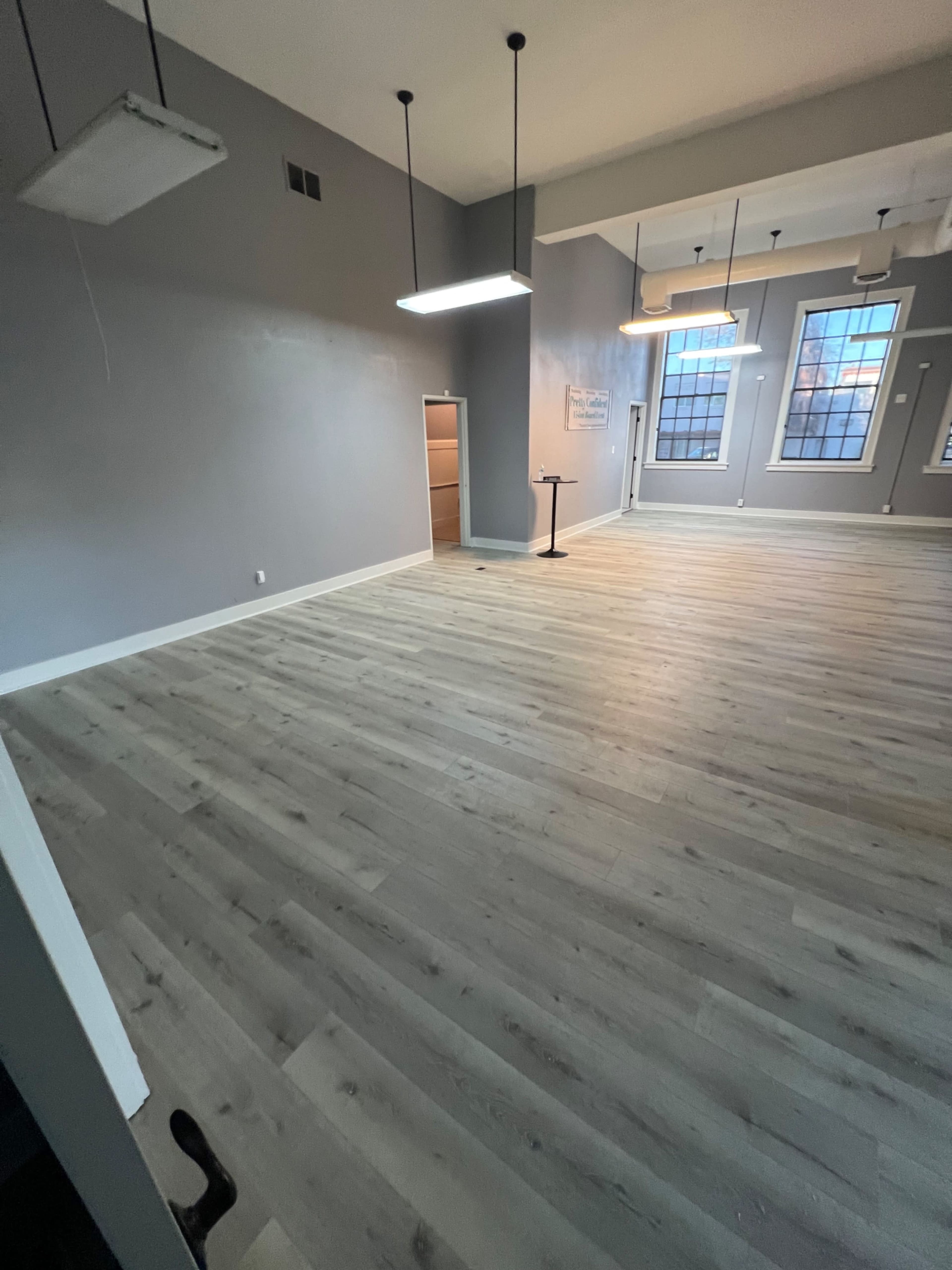 The image shows an empty room with gray walls, large windows, and light wood flooring.