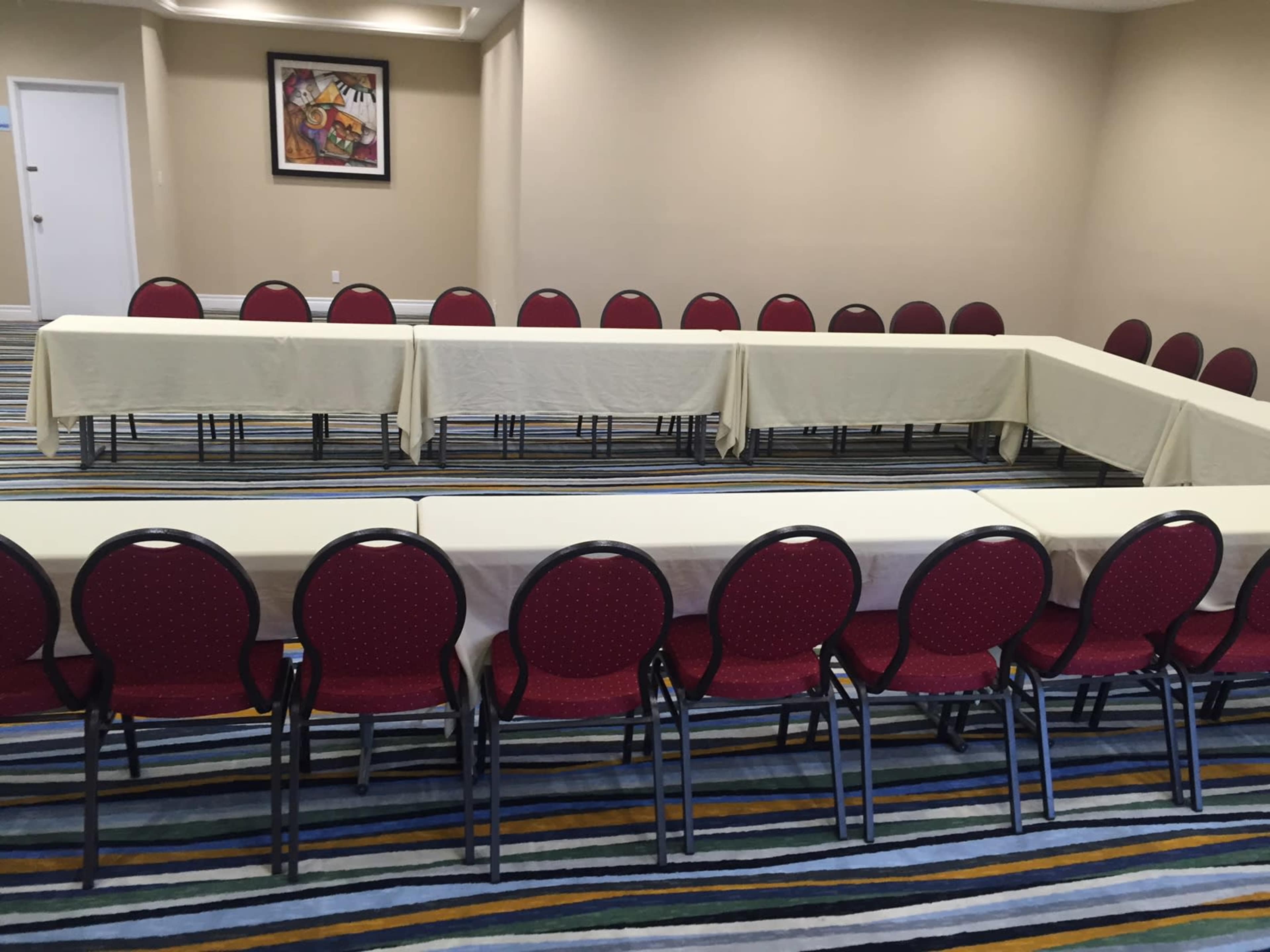 A conference room is set up with a rectangular arrangement of tables surrounded by red chairs on a striped carpet.