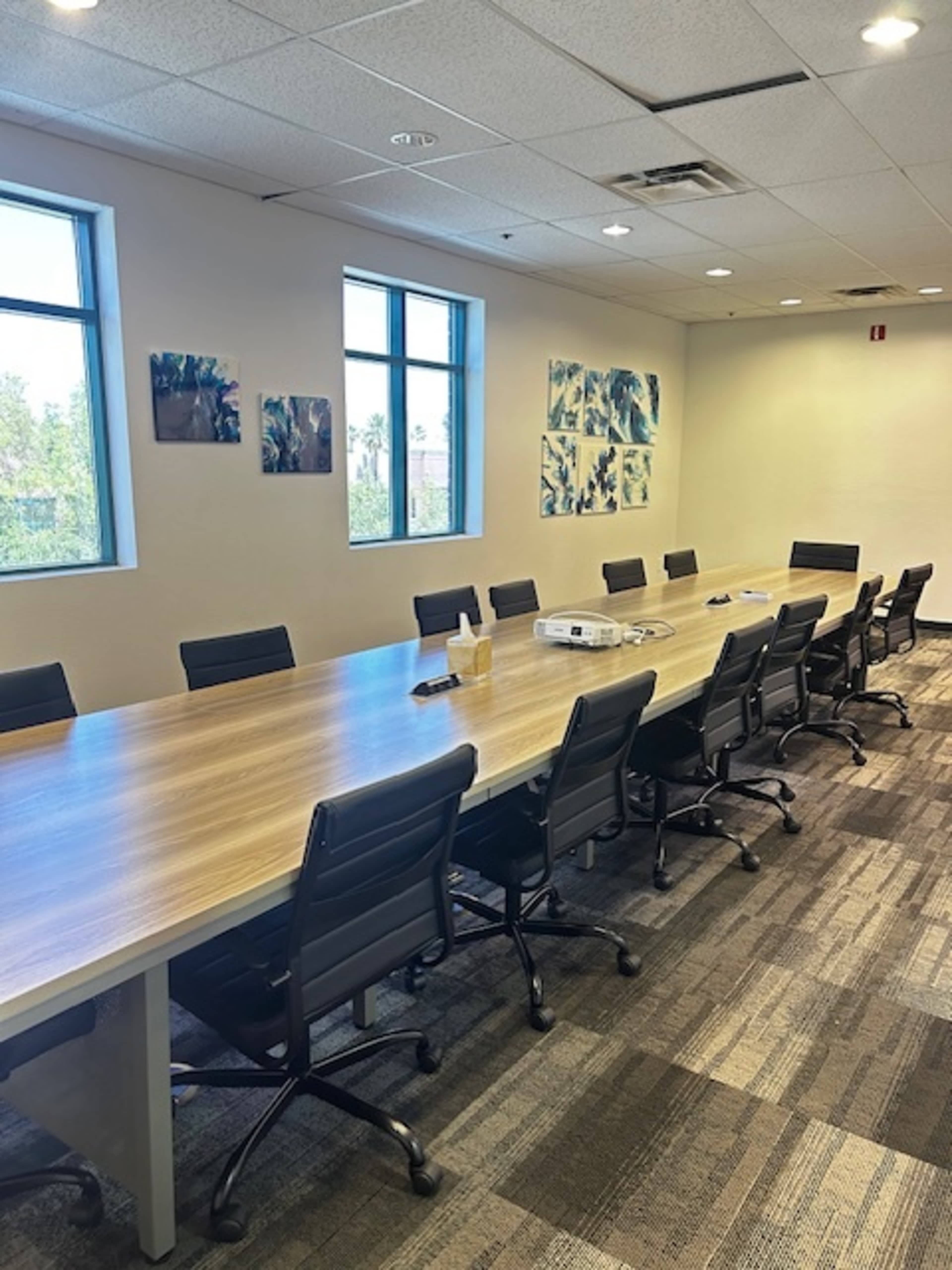 A long conference table with black chairs is set up in a brightly lit meeting room featuring windows and artwork on the walls.