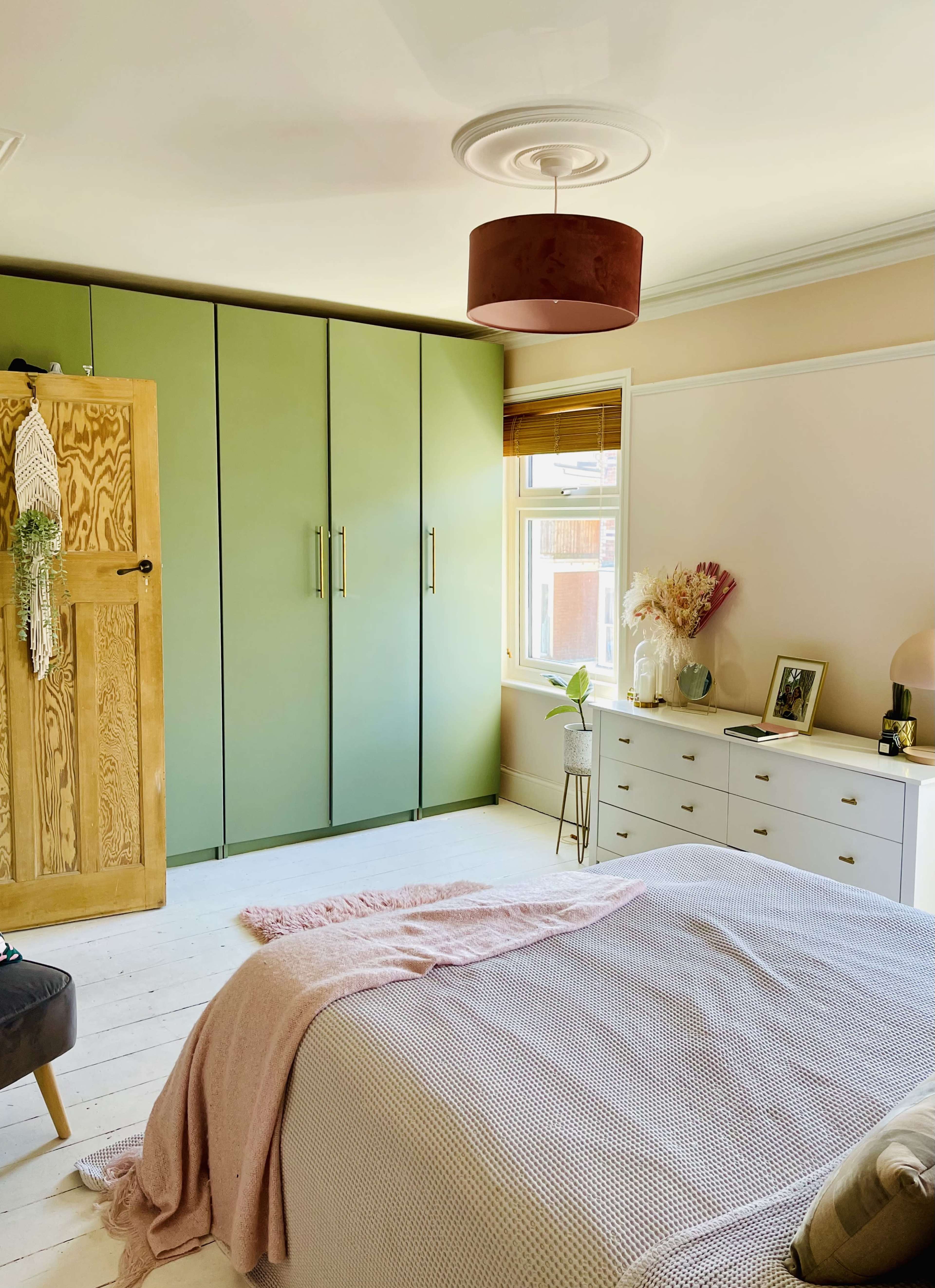 A well-organized bedroom featuring a light-colored bedspread, green wardrobes, a wooden door, and a white dresser with decorative items.