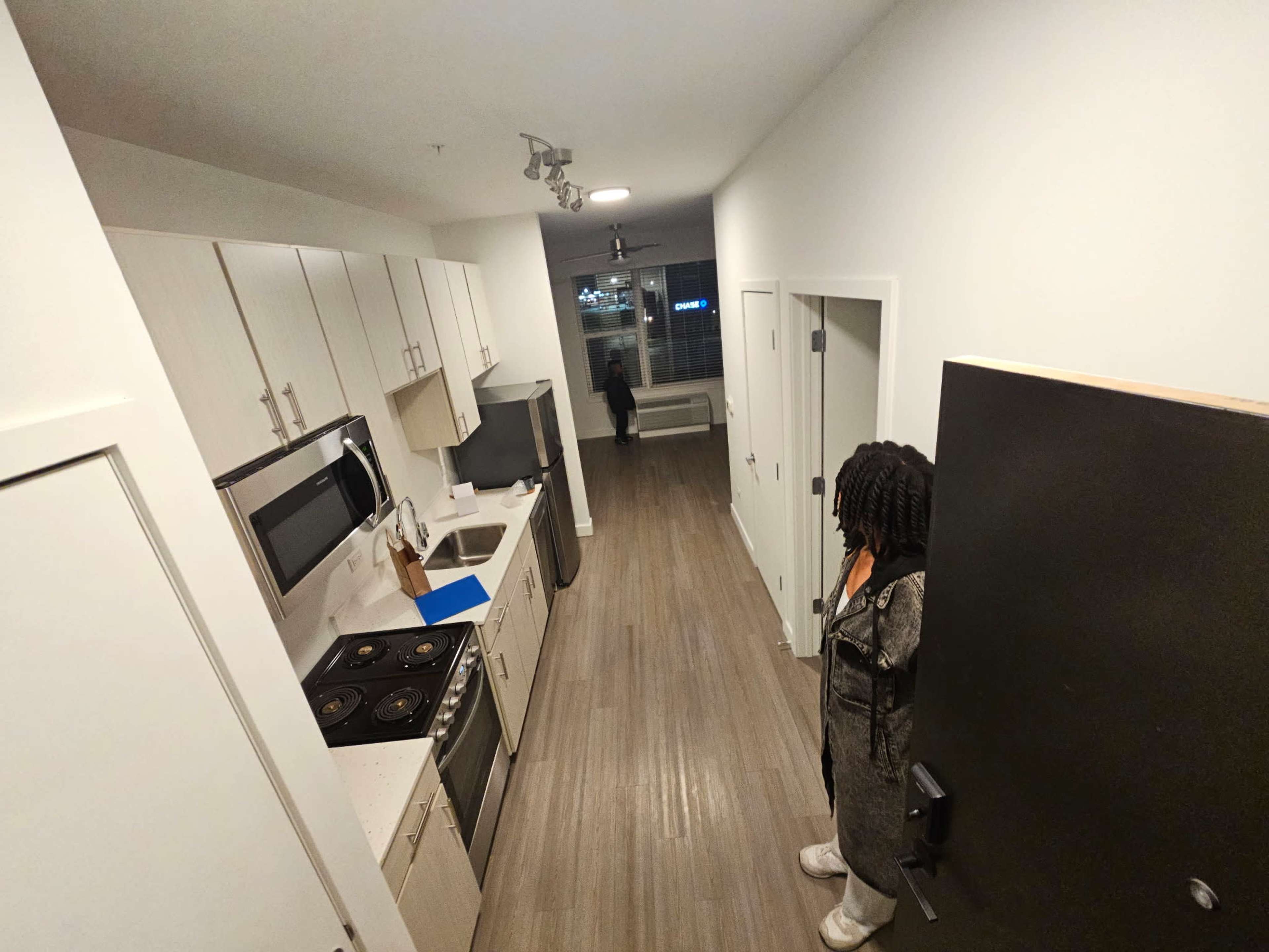 A view of a modern kitchen with white cabinetry and stainless steel appliances, leading into a hallway with a person standing near the entrance door.