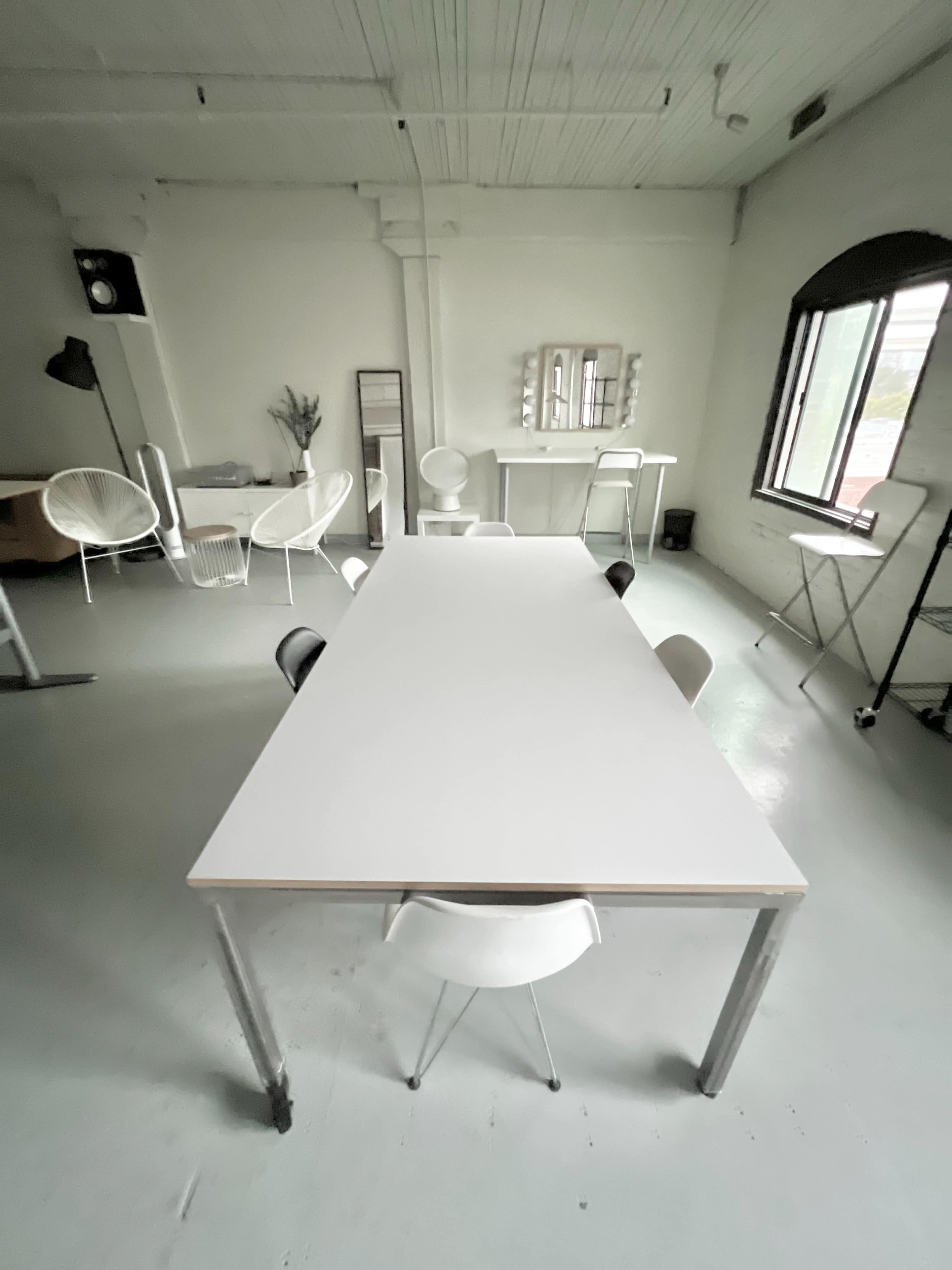A spacious, minimalist room features a long white table surrounded by chairs, with additional furniture and large windows in the background.