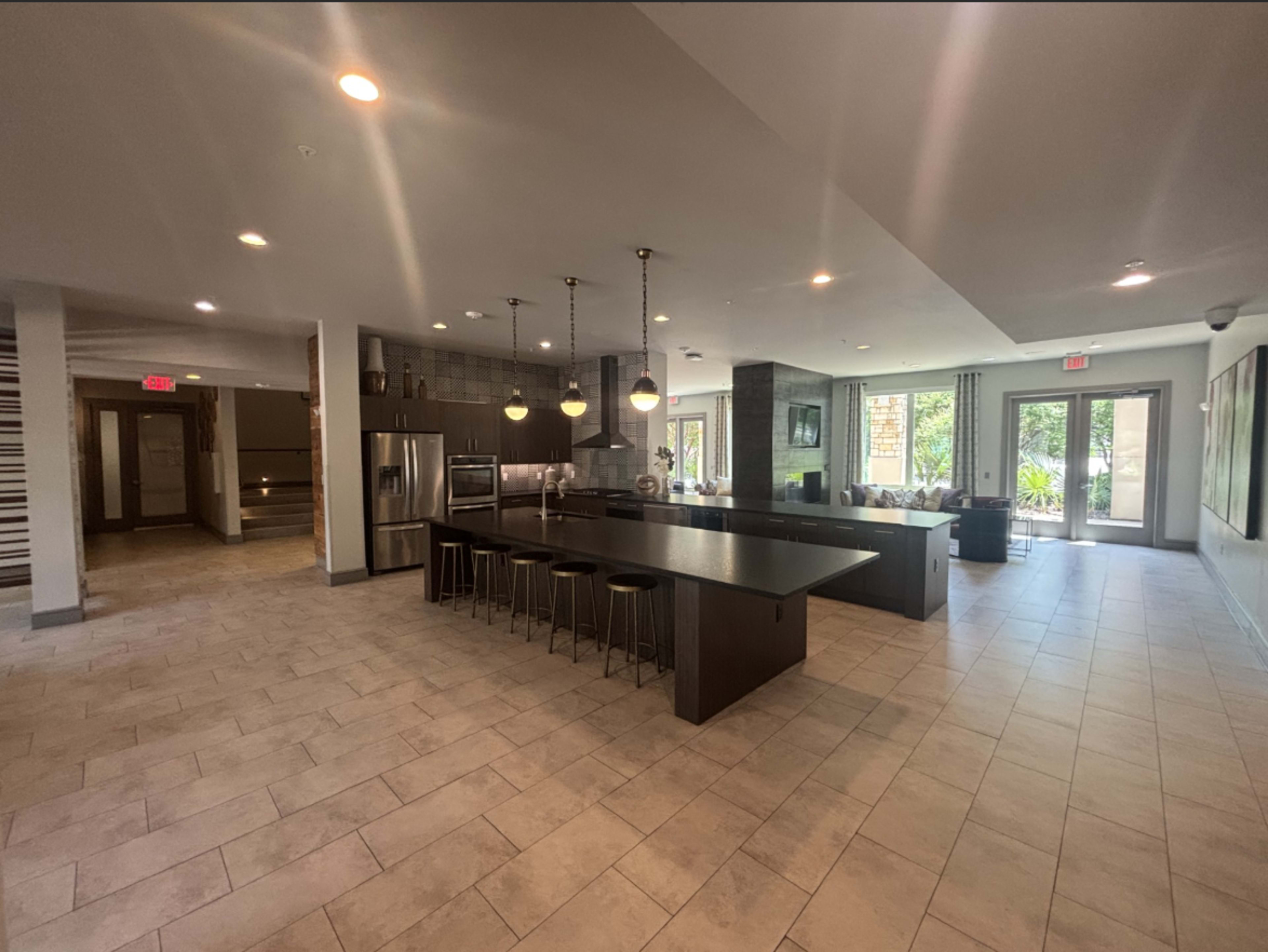 The image shows a modern kitchen and dining area with a large island and seating, surrounded by polished tile floors and large windows letting in natural light.