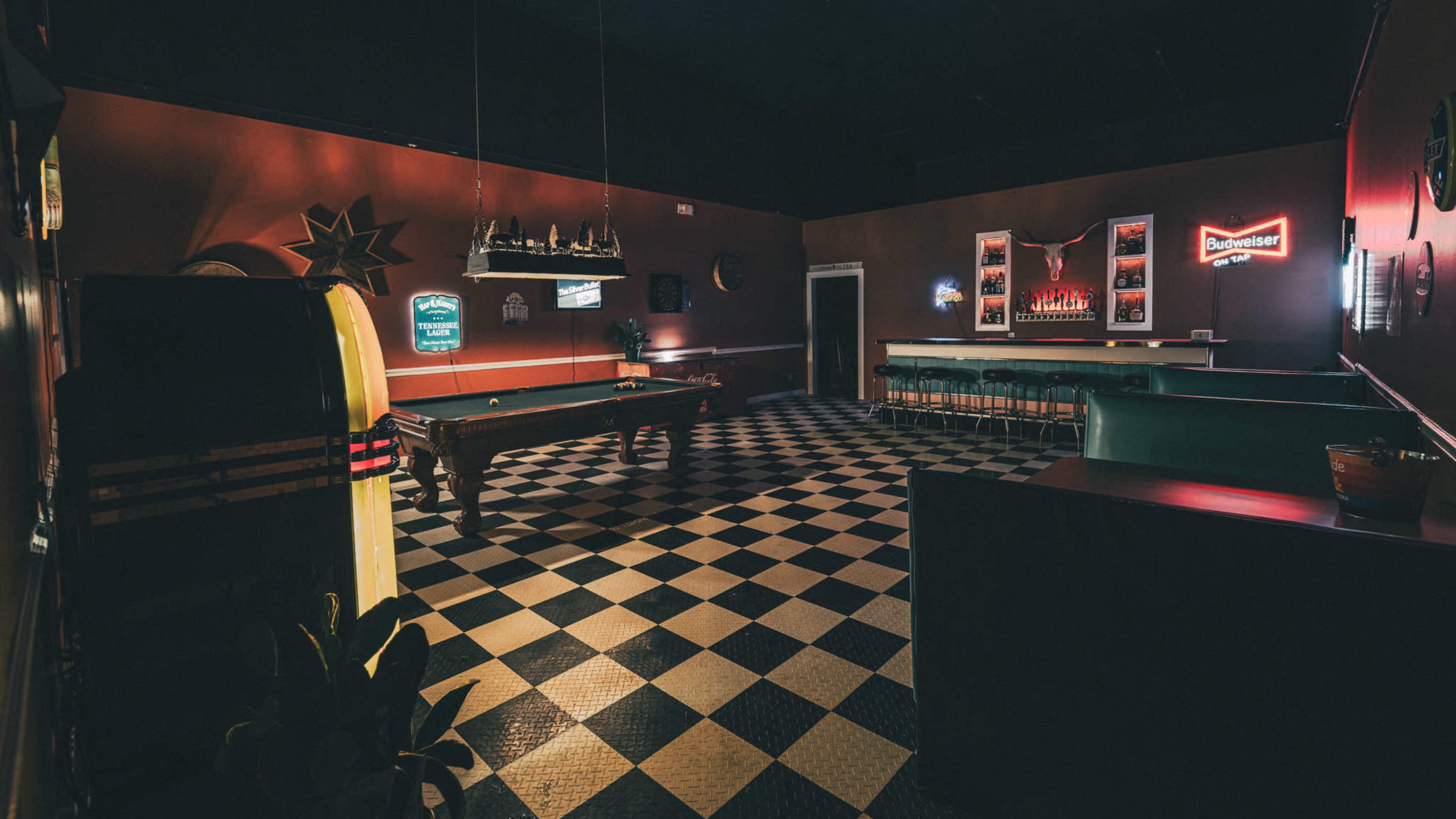 The image depicts a dimly lit game room featuring a pool table, a retro jukebox, and a bar counter with various neon signs on the walls.
