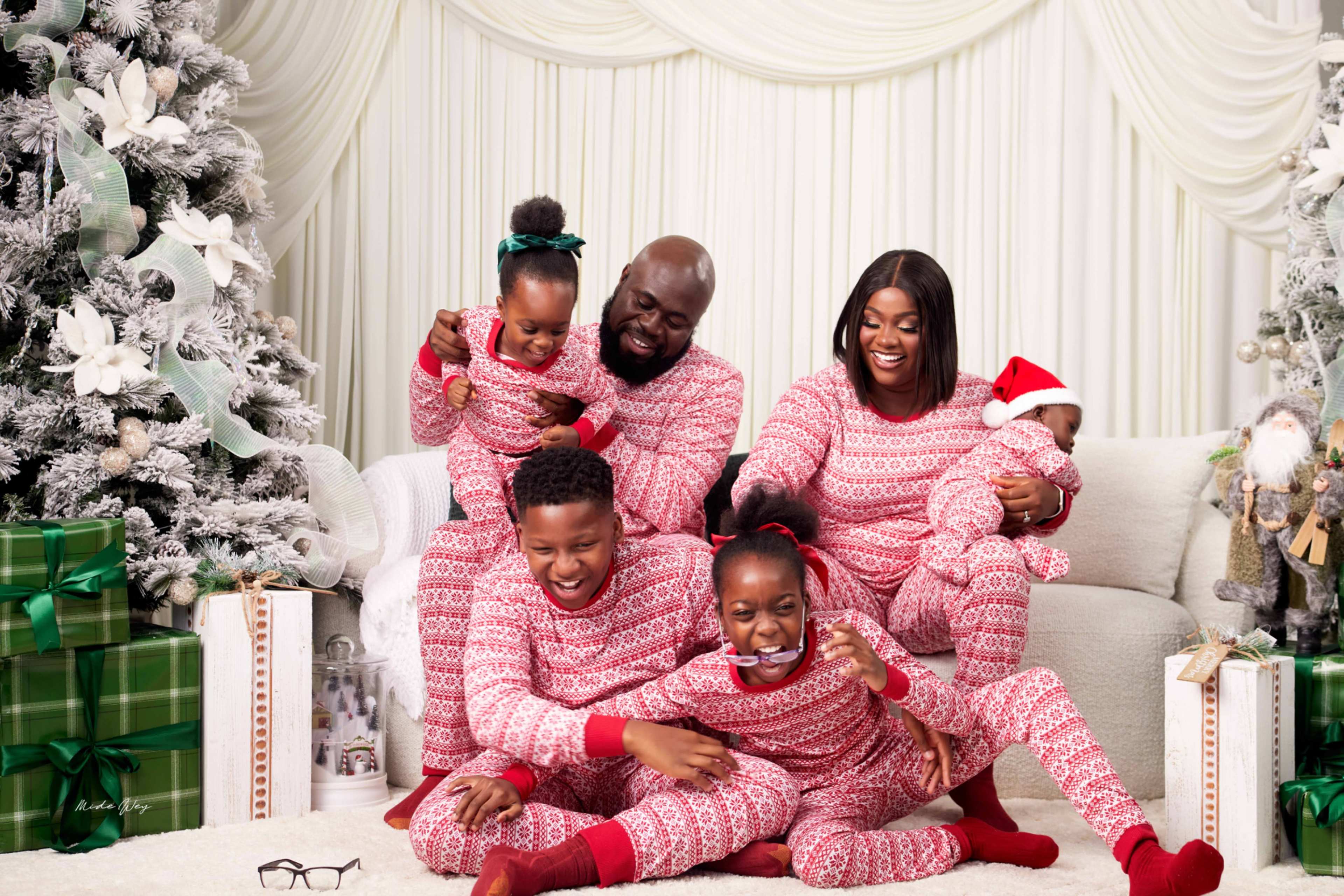 A family poses together on a couch, wearing matching red and white patterned pajamas, surrounded by Christmas decorations and gifts.