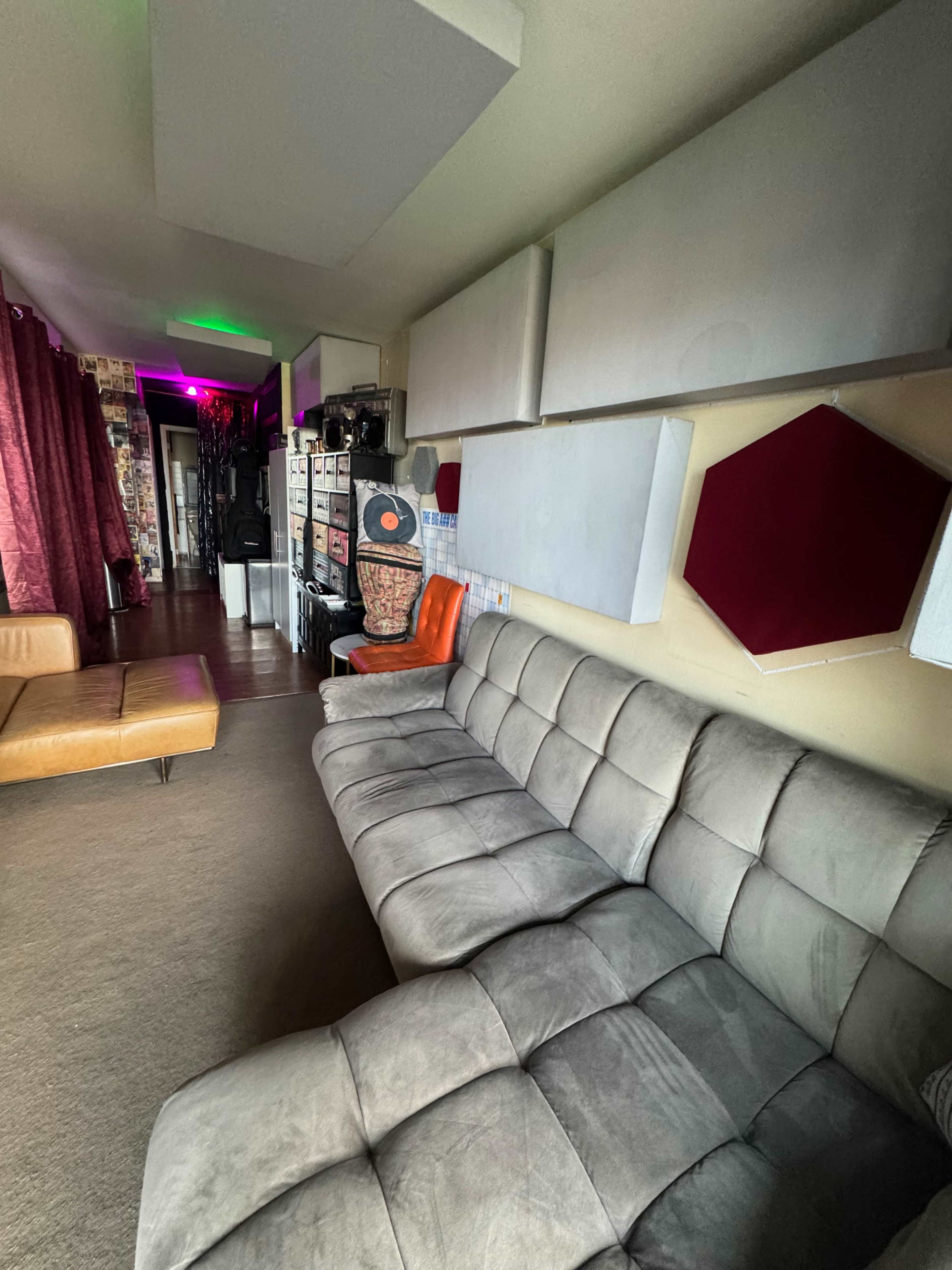 The image shows a room with a gray sectional sofa, a brown leather couch, and a wall featuring various soundproof panels and decor.