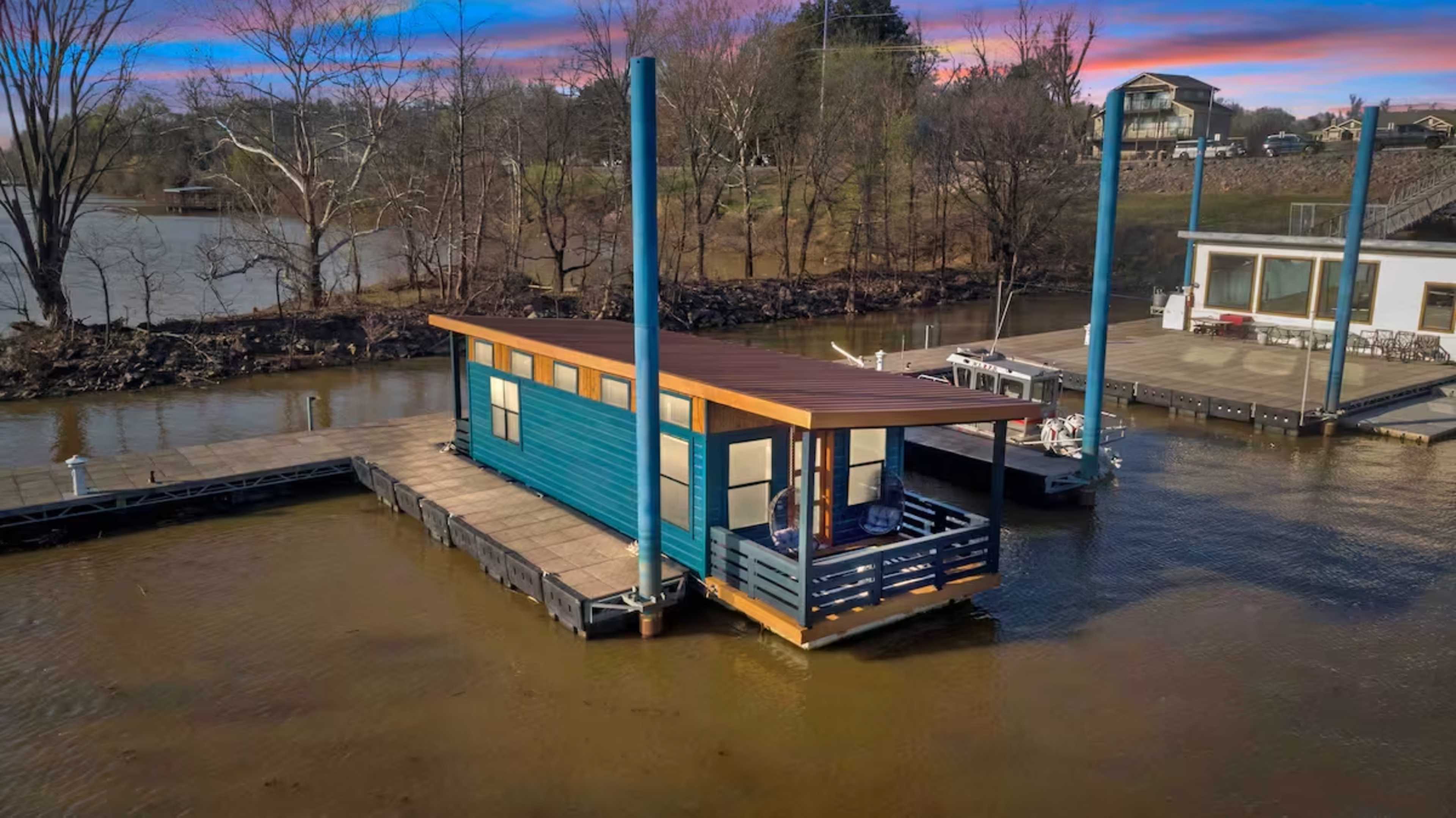 The image shows a colorful houseboat docked at a pier in calm waters, surrounded by trees and buildings in the background.