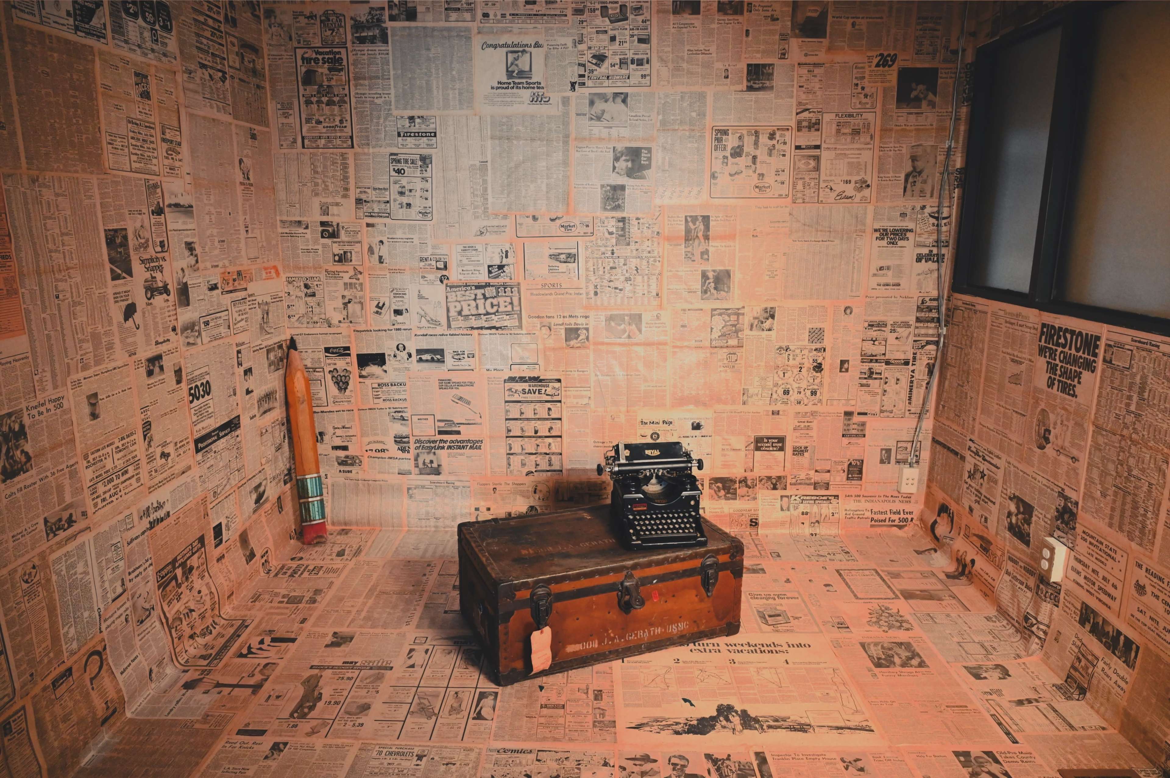 A small room where the walls, floor, and a vintage suitcase are covered in old newspapers, alongside a typewriter placed on top of the suitcase.