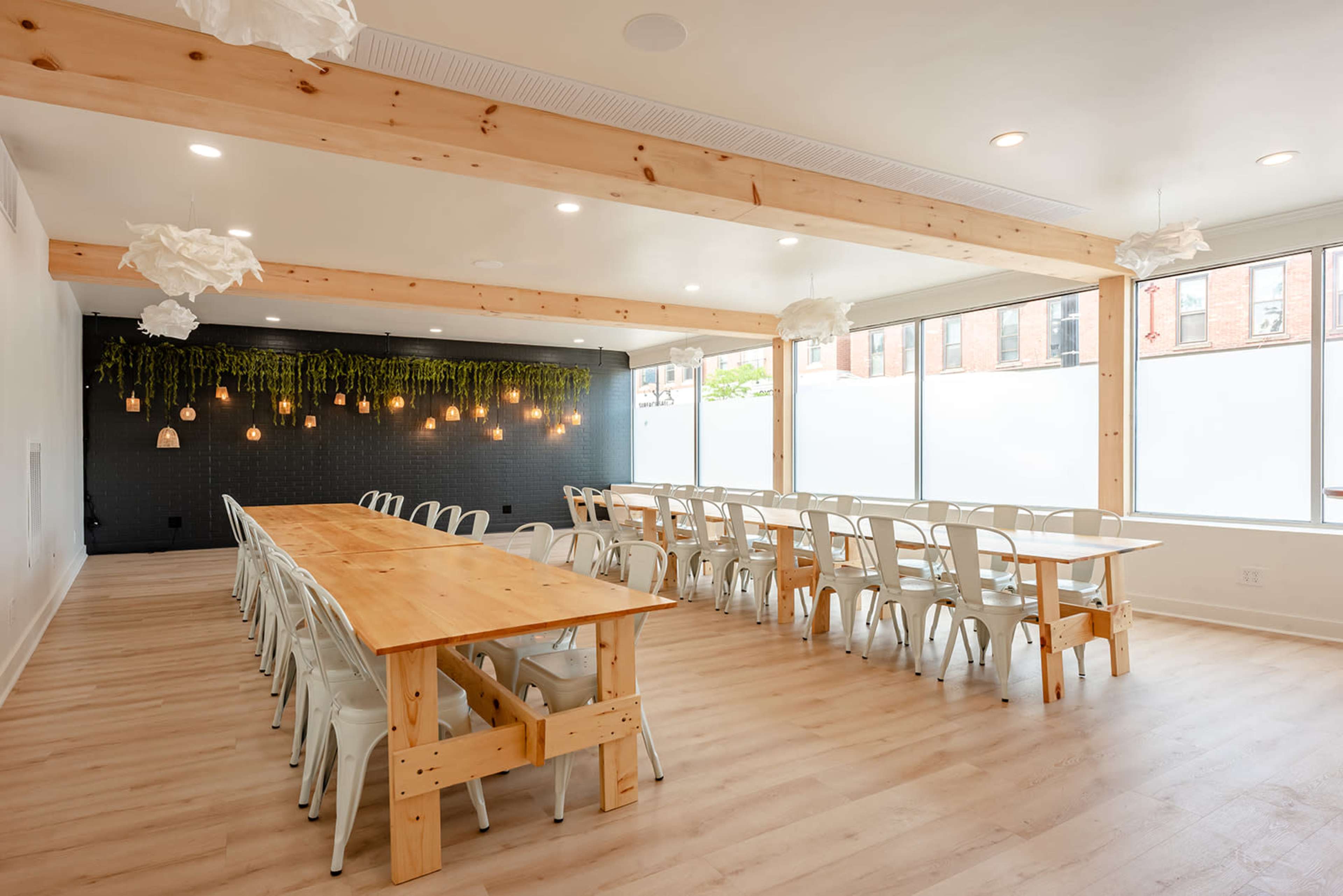 A spacious, well-lit dining area features long wooden tables arranged with white chairs and a green wall of hanging plants.