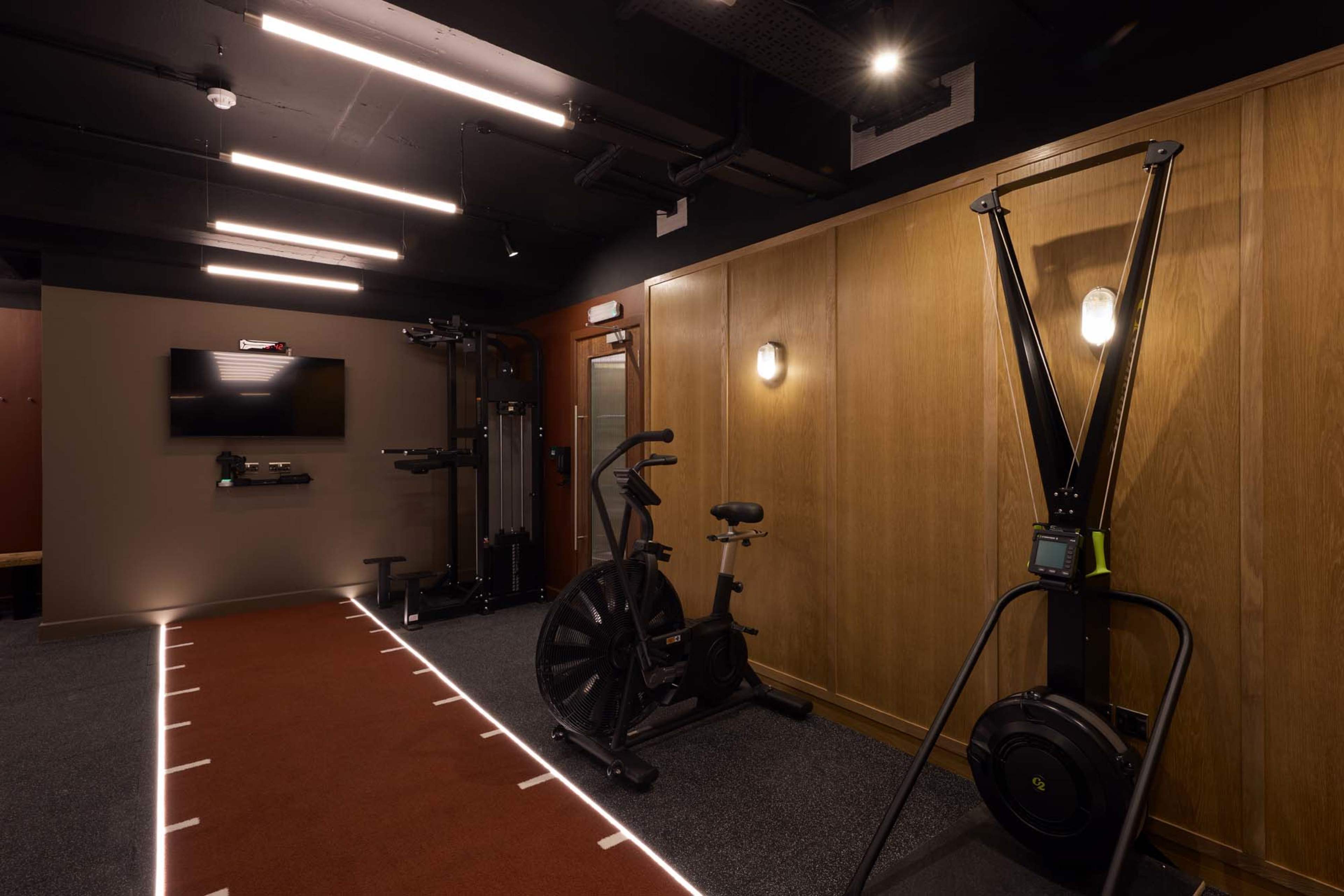 The image shows a modern gym area featuring various workout equipment, including an exercise bike, a rowing machine, and strength training apparatus, along with track lighting and wooden paneling.