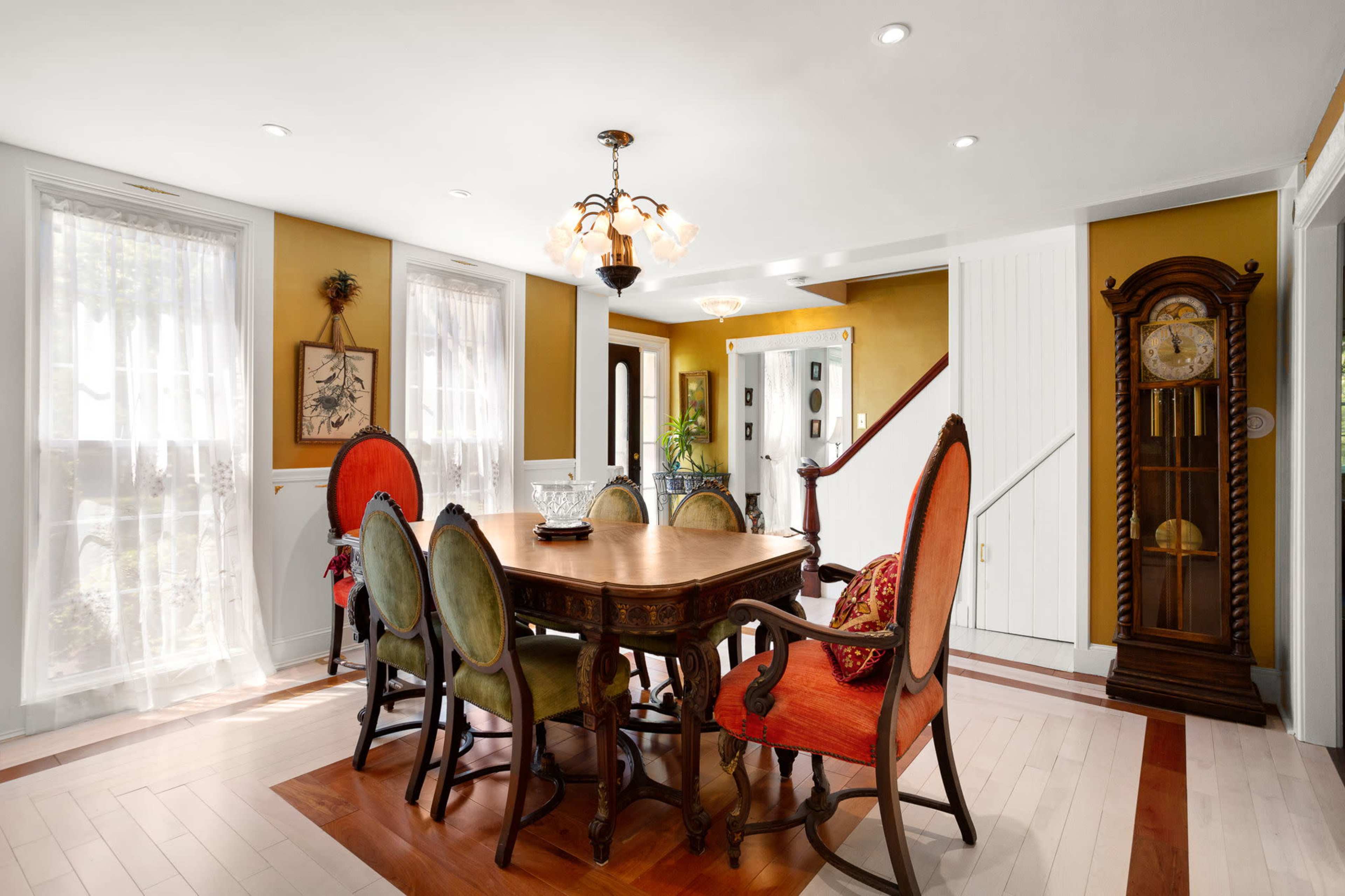 A dining area features a wooden table surrounded by six chairs, with a chandelier overhead and a clock positioned against the wall.
