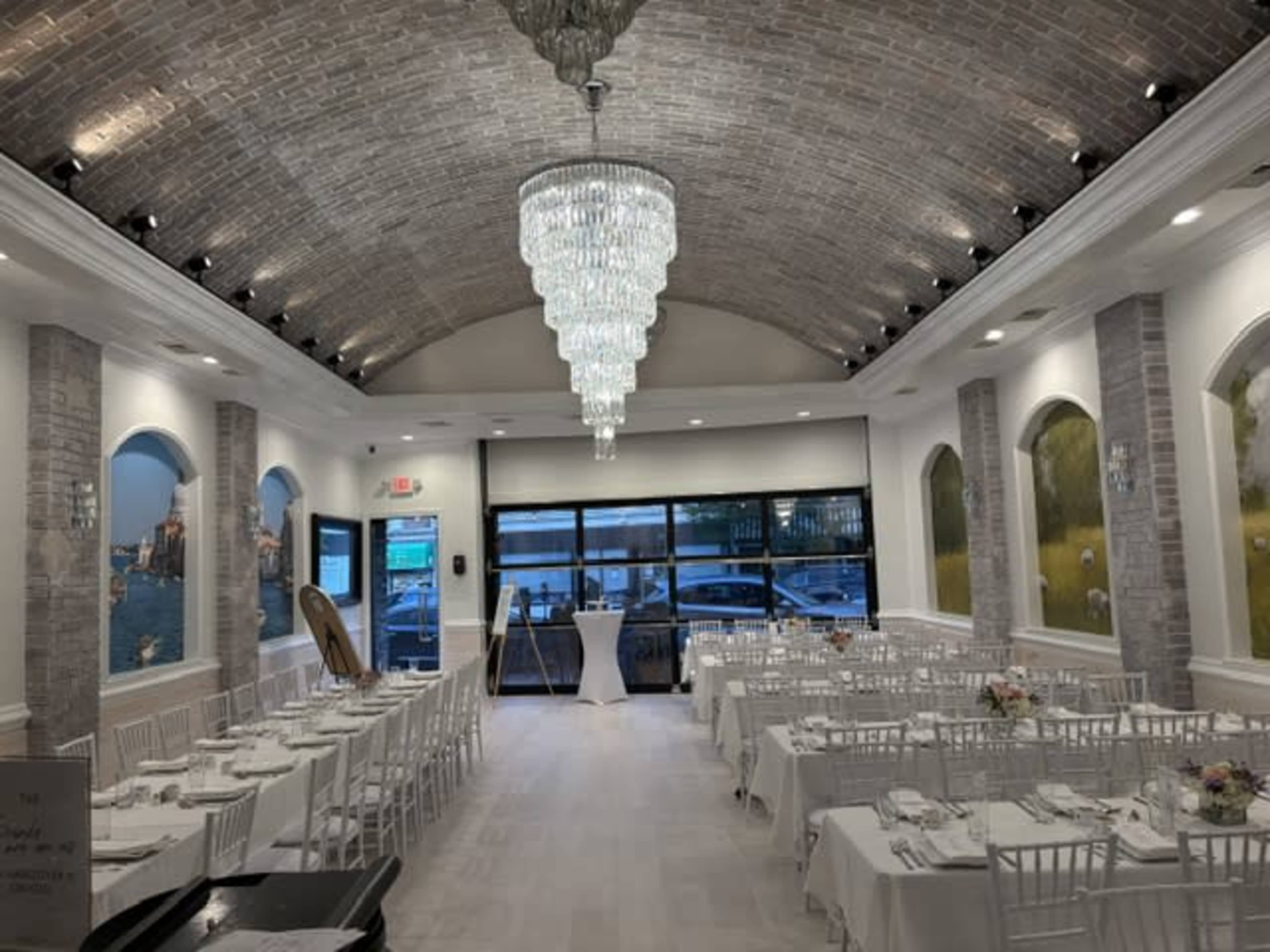 The image shows a well-decorated dining area with white tables set for a formal event, a chandelier hanging from the high ceiling, and large windows providing views outside.