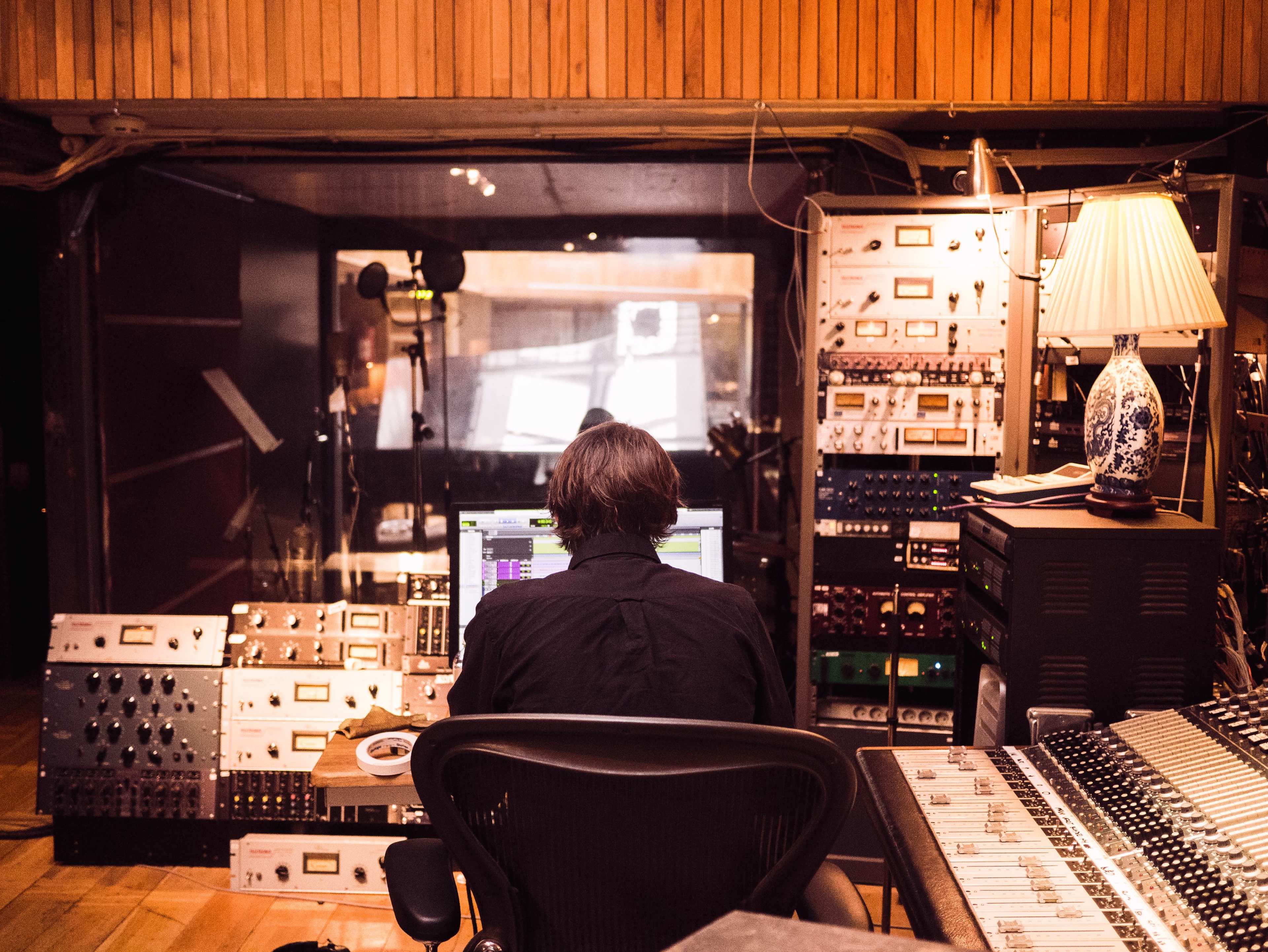 A person operates a digital audio workstation in a recording studio filled with audio equipment and a mixing console.